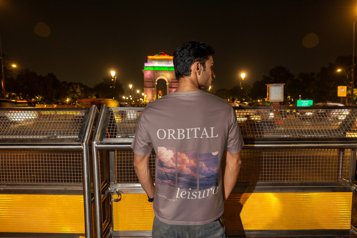 Stay for the Sky Garment-Dyed Tee in Espresso, worn by a man seen from behind on a bridge over the city at night, showing the large print on the back. Digital product mockup.
