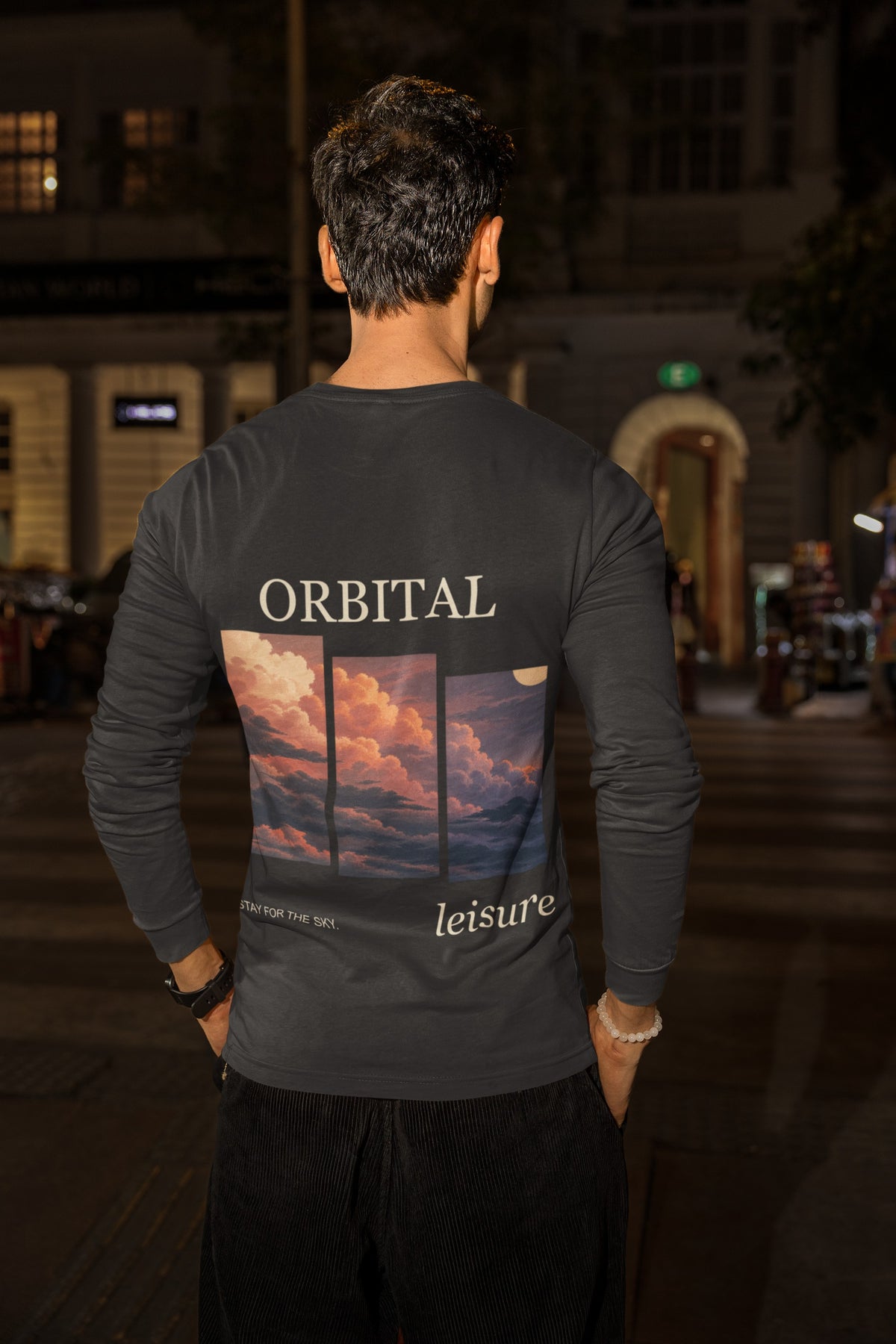 Stay for the Sky Garment-Dyed Long-Sleeve Tee in Black, worn by a man seen from behind on a city street at night, showing the large print on the back. Digital product mockup.