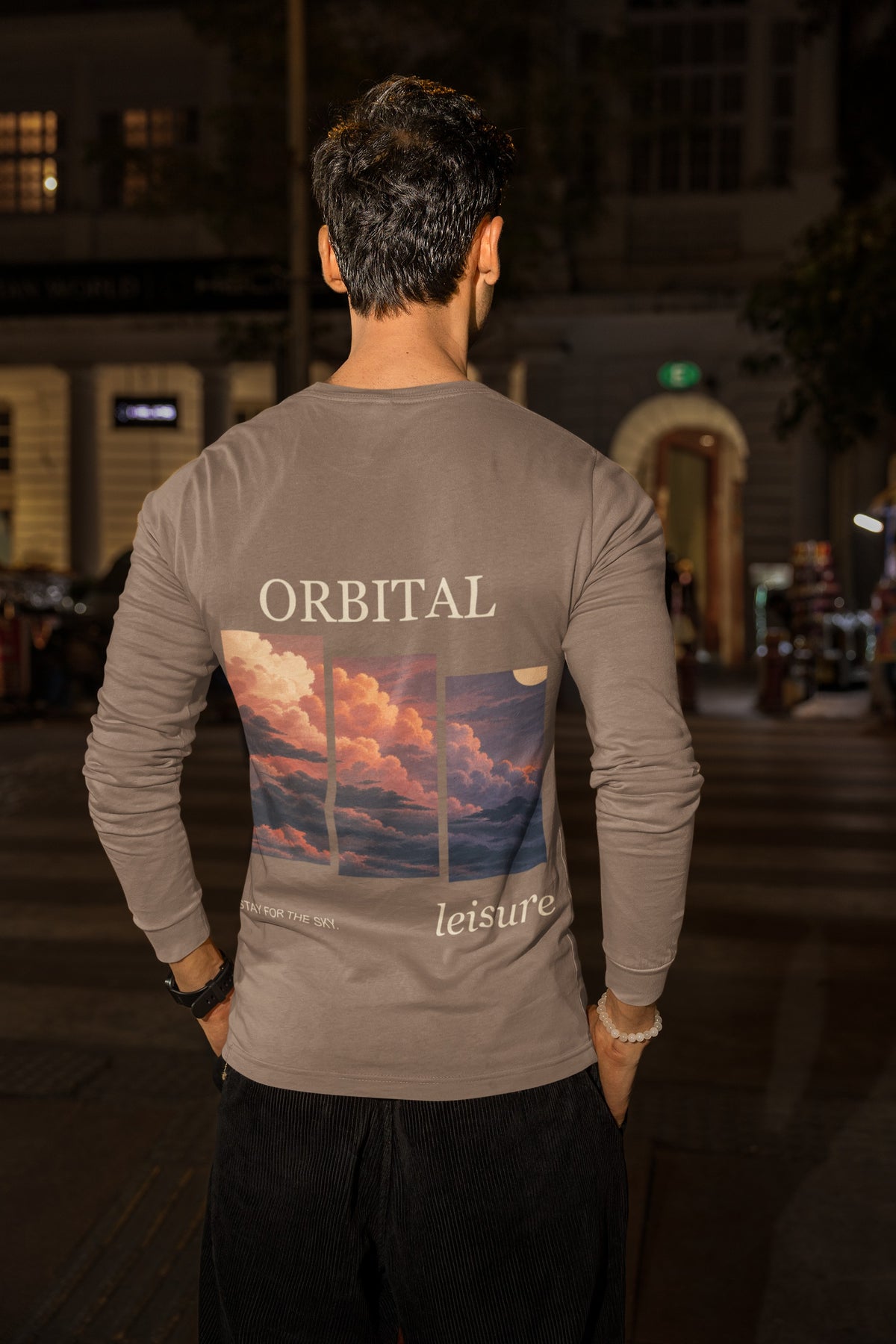 Stay for the Sky Garment-Dyed Long-Sleeve Tee in Espresso, worn by a man seen from behind on a city street at night, showing the large print on the back. Digital product mockup.