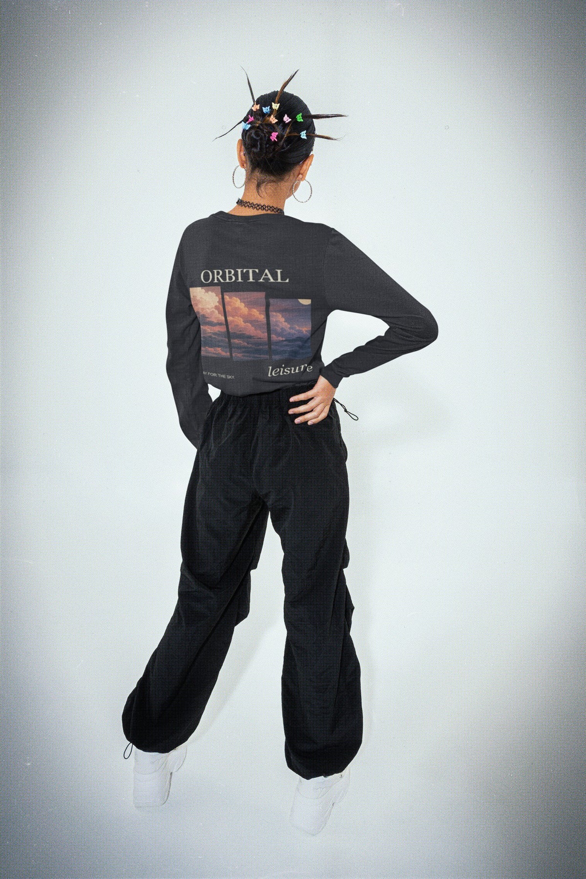 Stay for the Sky Garment-Dyed Long-Sleeve Tee in Black, worn by a woman seen from behind in a studio, showing the large print on the back. Digital product mockup.