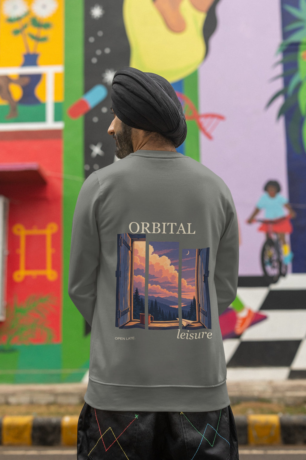 Open Late Garment-Dyed Crewneck in Pepper, worn by a man seen from behind in a turban by a painted mural, showing the large print on the back. Digital product mockup.