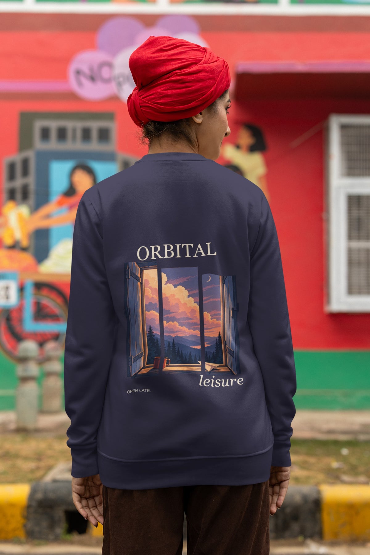 Open Late Garment-Dyed Crewneck in True Navy, worn by a woman seen from behind in a turban by a painted mural, showing the large print on the back. Digital product mockup.