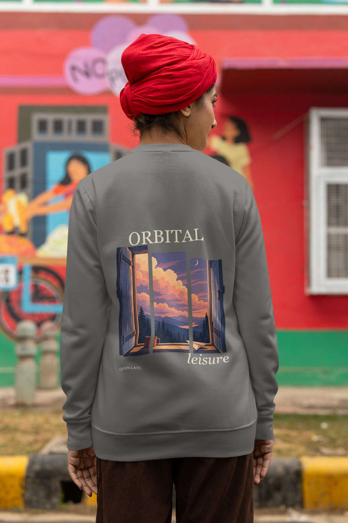 Open Late Garment-Dyed Crewneck in Pepper, worn by a woman seen from behind in a turban by a painted mural, showing the large print on the back. Digital product mockup.