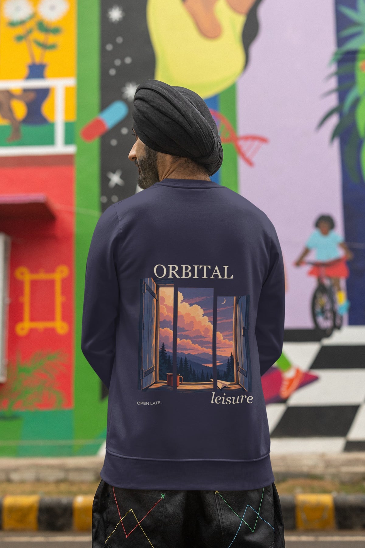Open Late Garment-Dyed Crewneck in True Navy, worn by a man seen from behind in a turban by a painted mural, showing the large print on the back. Digital product mockup.