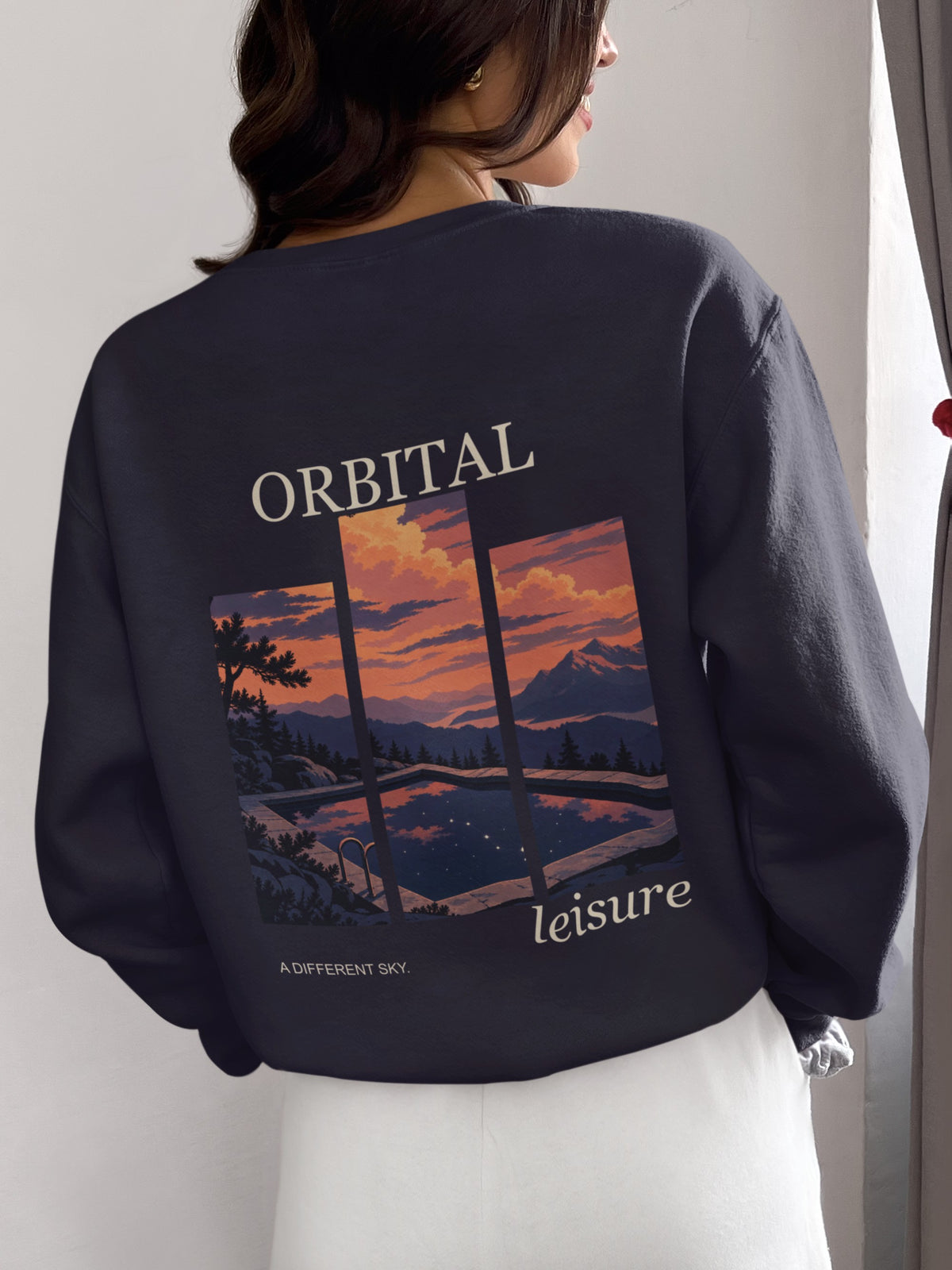 A Different Sky Garment-Dyed Crewneck in True Navy, worn by a woman seen from behind at home, showing the large print on the back. Digital product mockup.