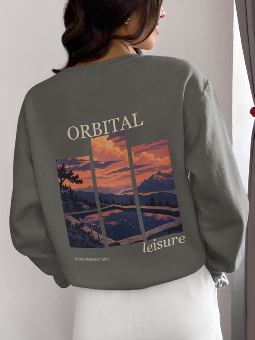 A Different Sky Garment-Dyed Crewneck in Pepper, worn by a woman seen from behind at home, showing the large print on the back. Digital product mockup.