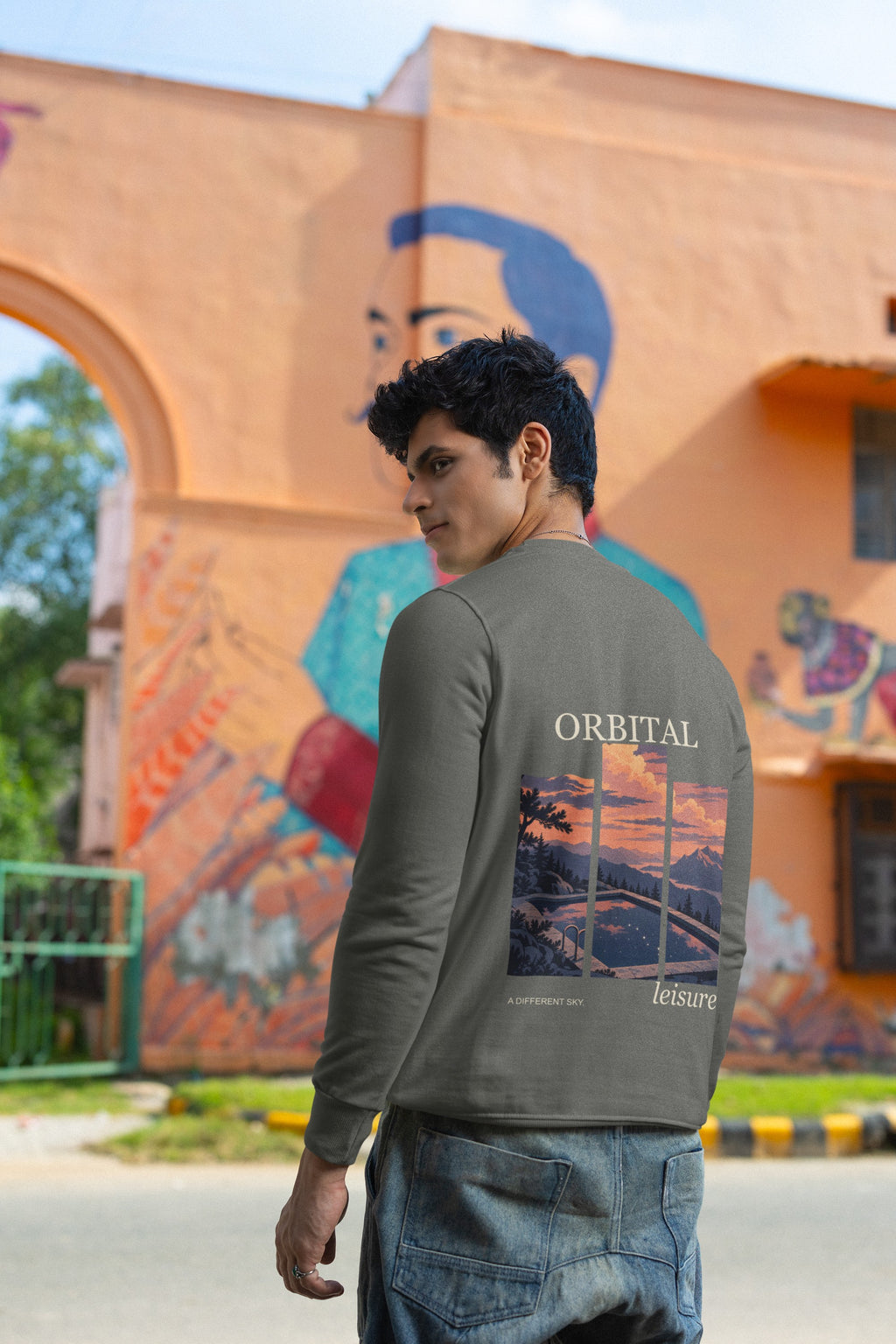 A Different Sky Garment-Dyed Crewneck in Pepper, worn by a man seen from behind by a painted wall in the city, showing the large print on the back. Digital product mockup.
