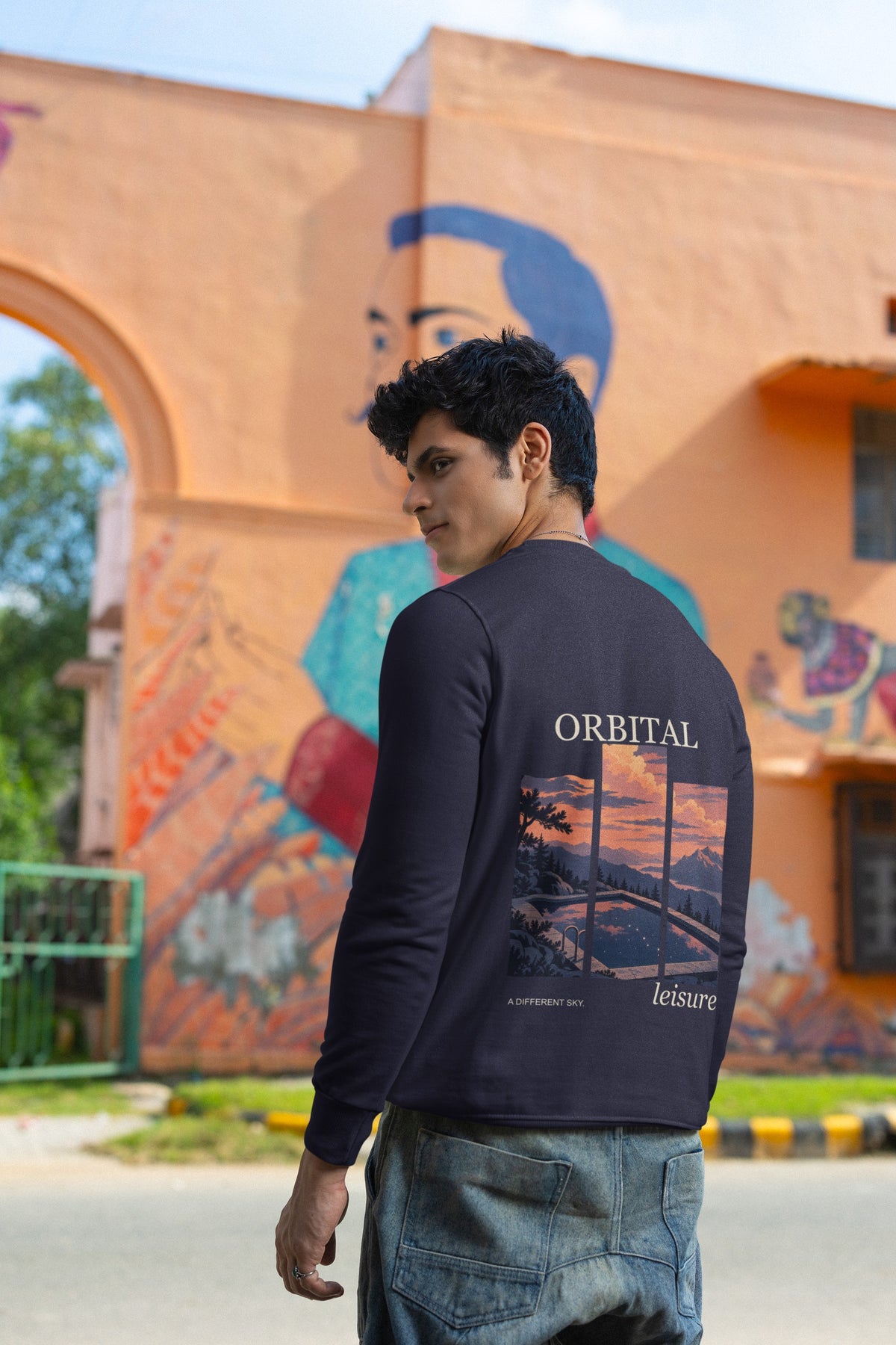 A Different Sky Garment-Dyed Crewneck in True Navy, worn by a man seen from behind by a painted wall in the city, showing the large print on the back. Digital product mockup.