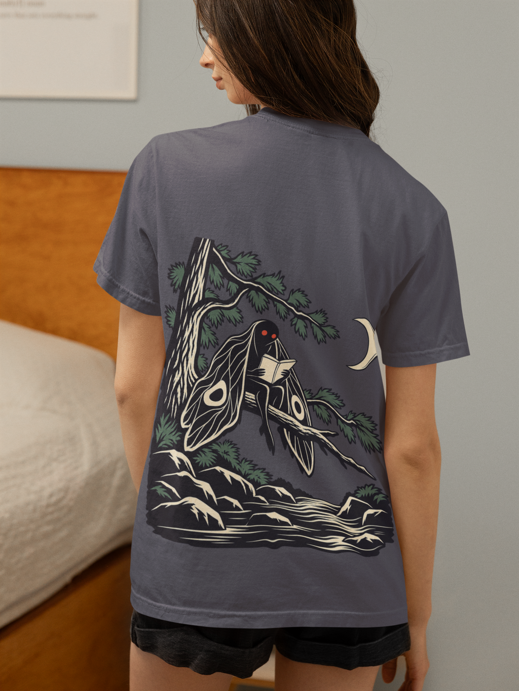Night Library Garment-Dyed Tee in Navy, worn by a woman seen from behind in a bedroom at home, showing the large print on the back. Digital product mockup.