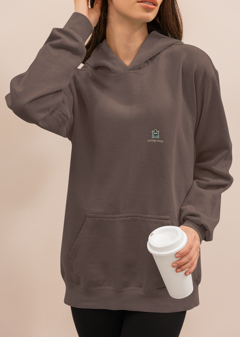 Night Library Heavyweight Hoodie in Brown, worn by a woman at home with a coffee cup, with the Night Library logo and portage.camp on the left chest. Digital product mockup.