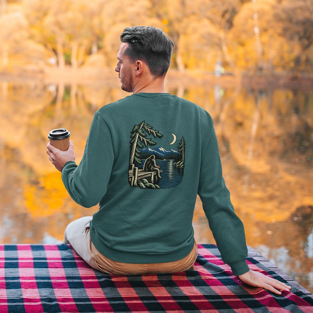 Quiet Hours Garment-Dyed Crewneck in Blue Spruce, worn by a man seen from behind seated by a lake in autumn, showing the large print on the back. Digital product mockup.