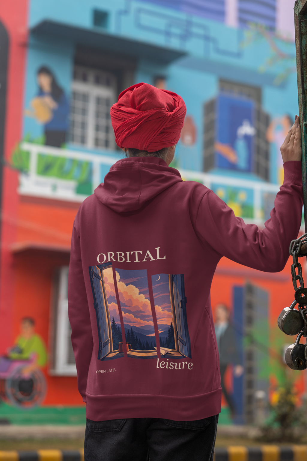 Open Late Midweight Hoodie in Maroon, worn by a woman seen from behind in a turban by a painted mural, showing the large print on the back. Digital product mockup.