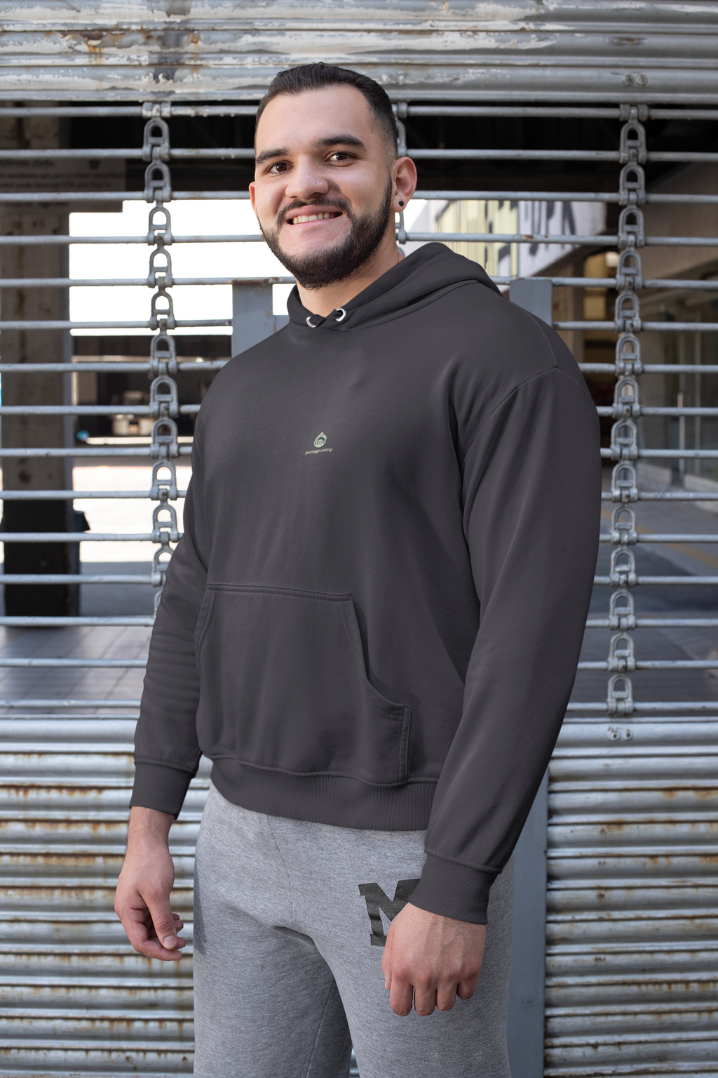 Rain Check Midweight Hoodie in Black, worn by a man against a metal fence, with the Rain Check logo and portage.camp on the chest. Digital product mockup.