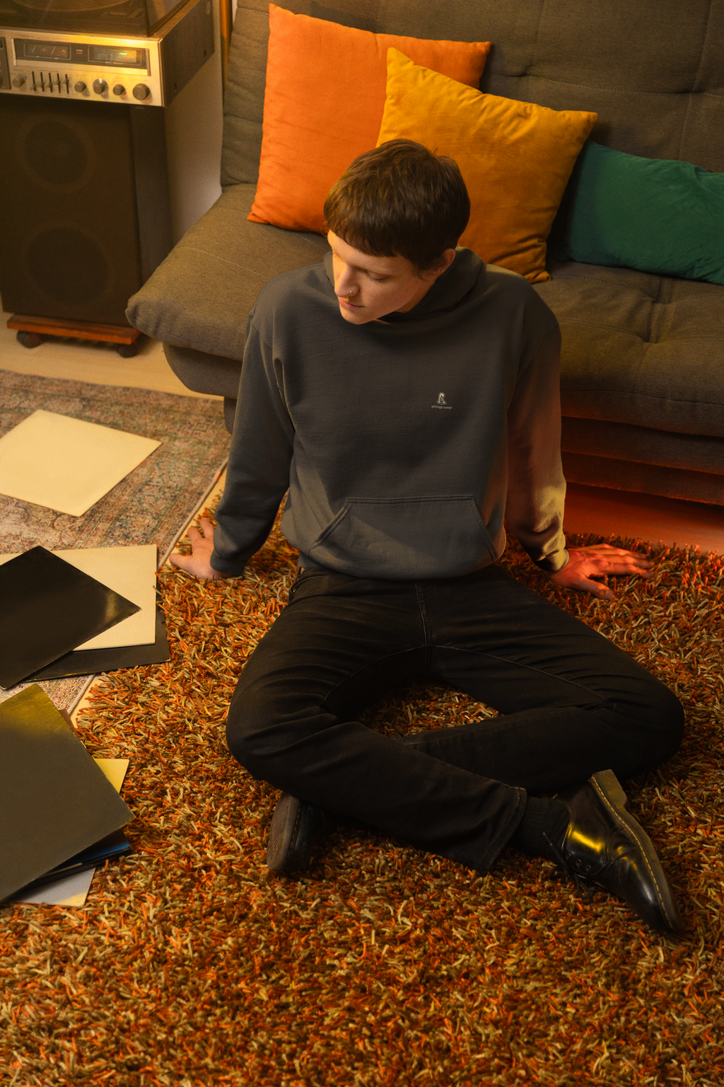 Small Hours Heavyweight Hoodie in Black, worn by a man seated on the floor among records, with the Small Hours logo and portage.camp on the left chest. Digital product mockup.