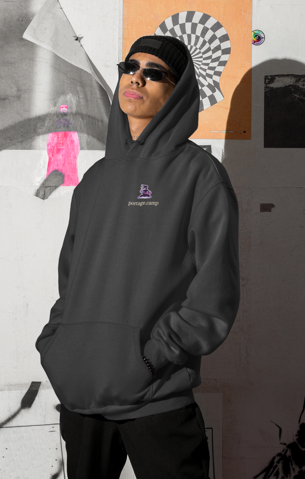 Orbit Keeper Heavyweight Hoodie in Black, worn by a man by a wall of posters, with the Orbit Keeper logo and portage.camp on the left chest. Digital product mockup.