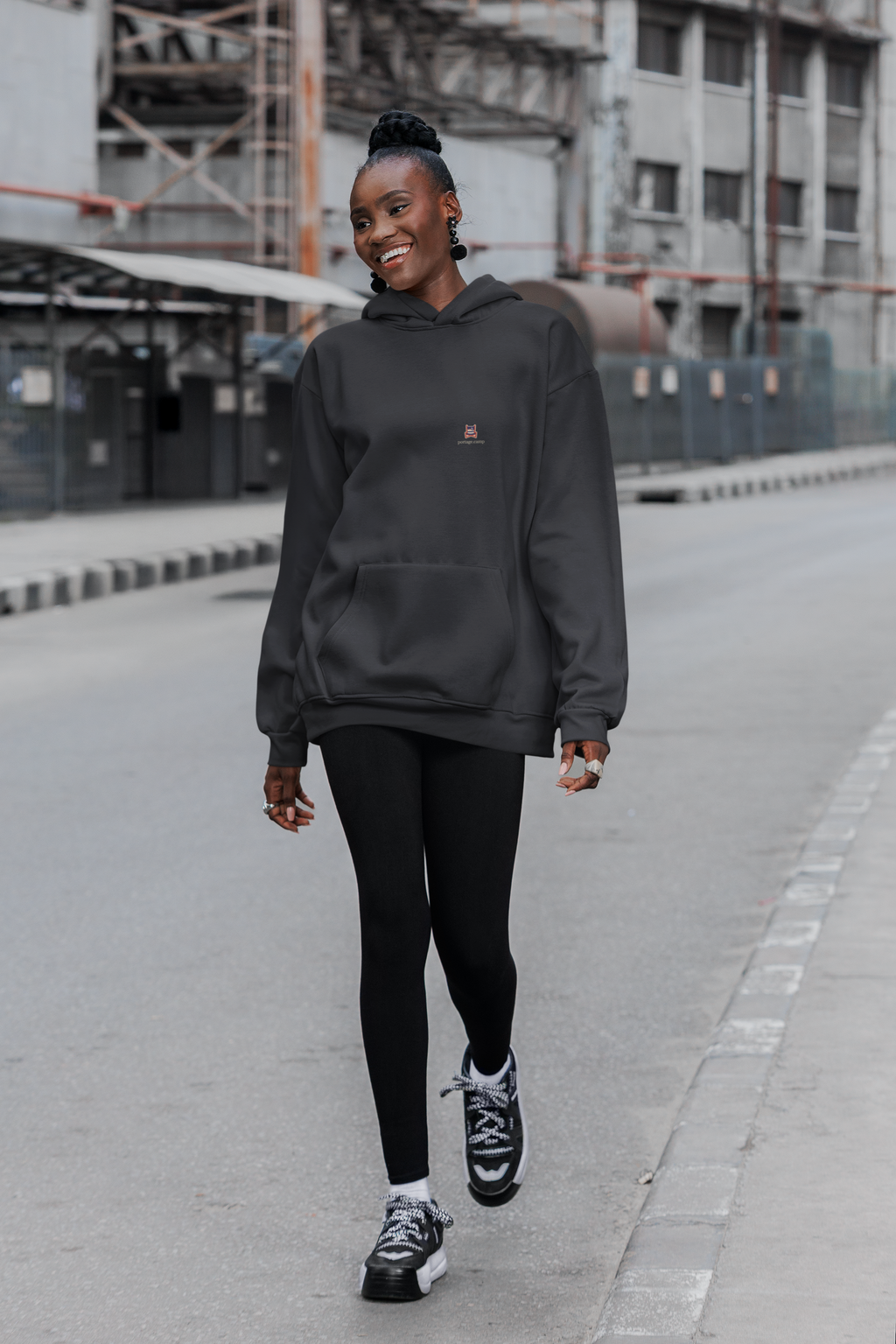 Edge of Evening Heavyweight Hoodie in Black, worn by a woman walking down a city street, with the Edge of Evening logo and portage.camp on the left chest. Digital product mockup.