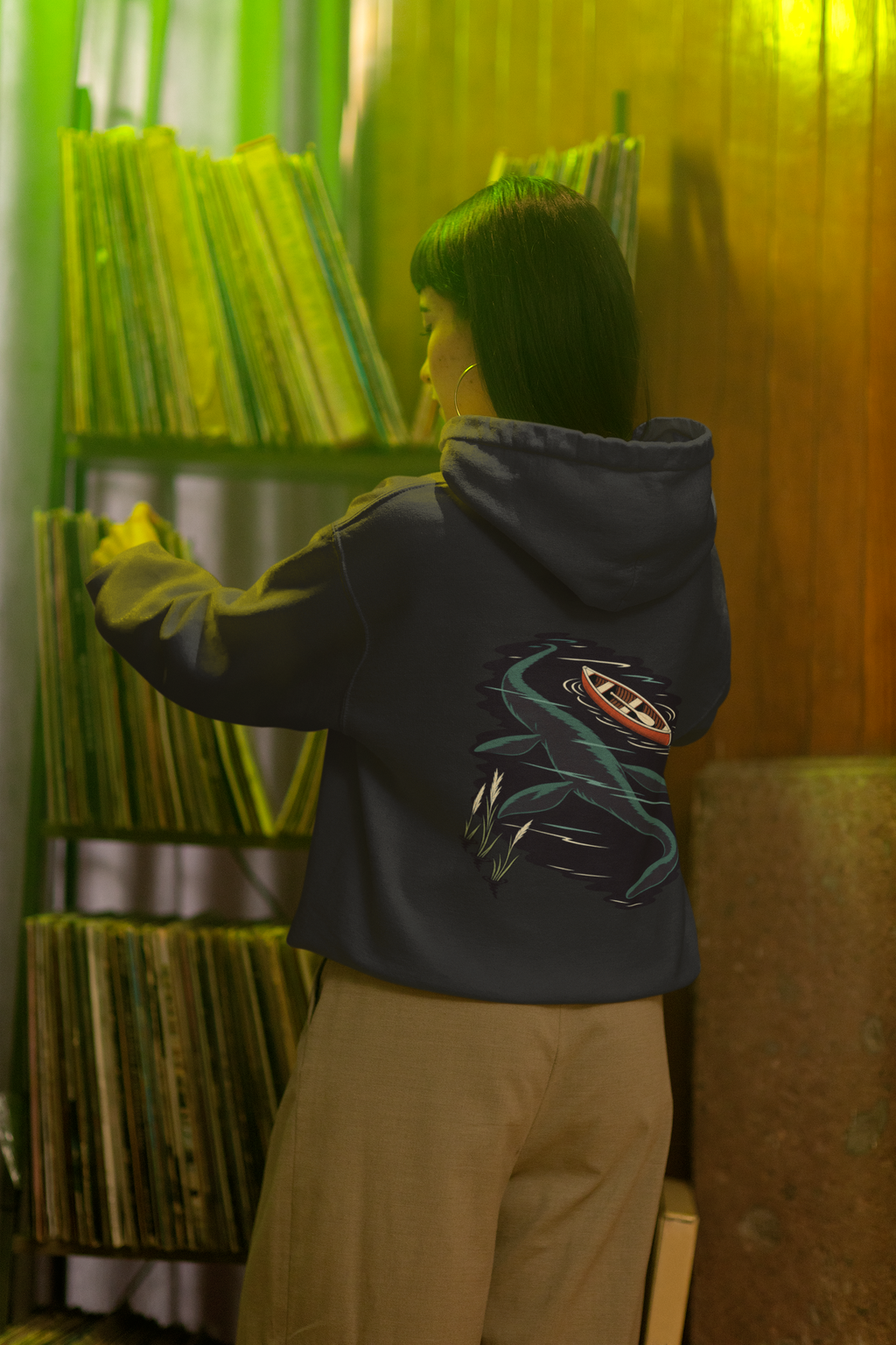 Seen Briefly Heavyweight Hoodie in Black, worn by a woman seen from behind in a vintage record shop, showing the large print on the back. Digital product mockup.