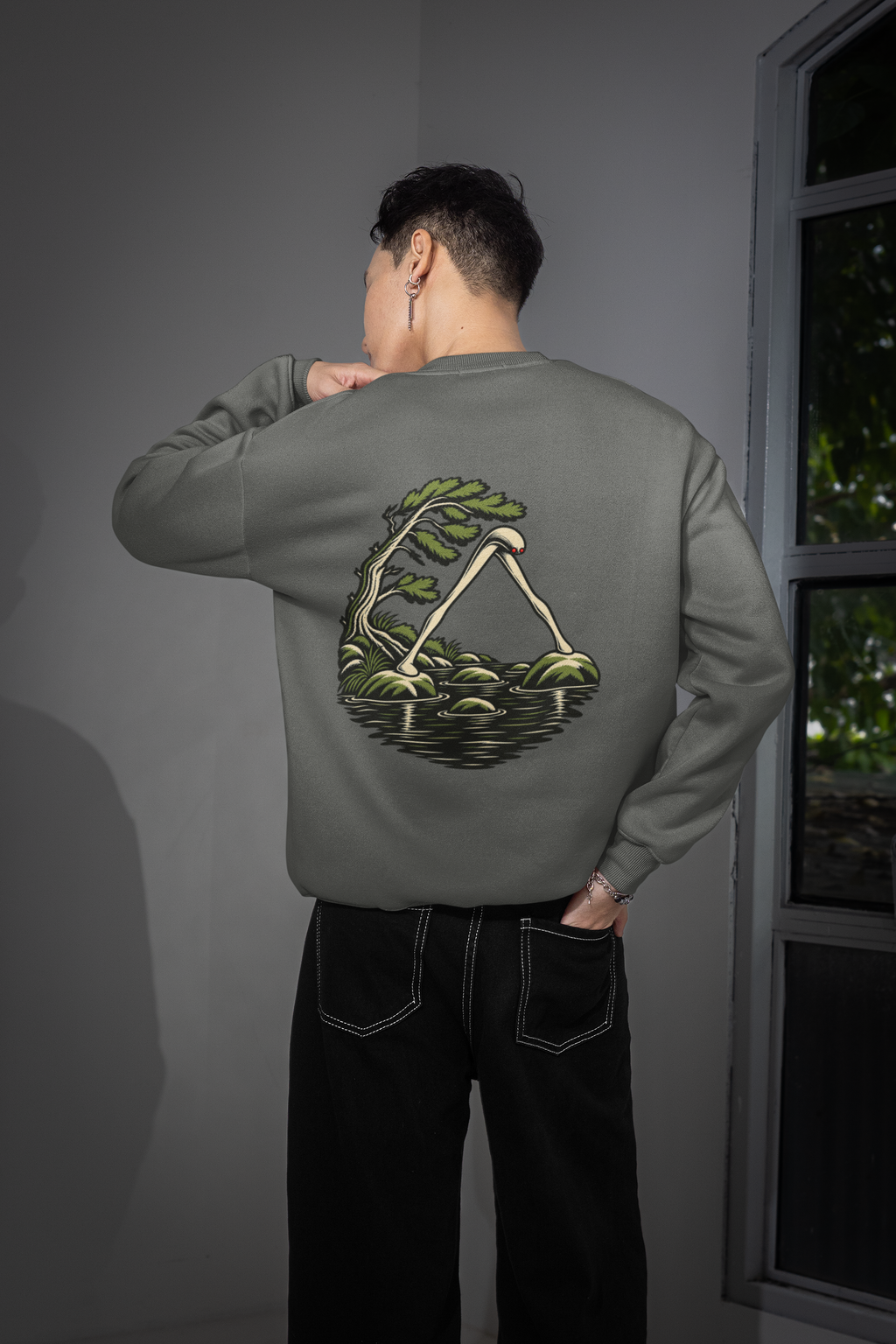 Small Hours Garment-Dyed Crewneck in Pepper, worn by a man seen from behind in a room by a window, showing the large print on the back. Digital product mockup.