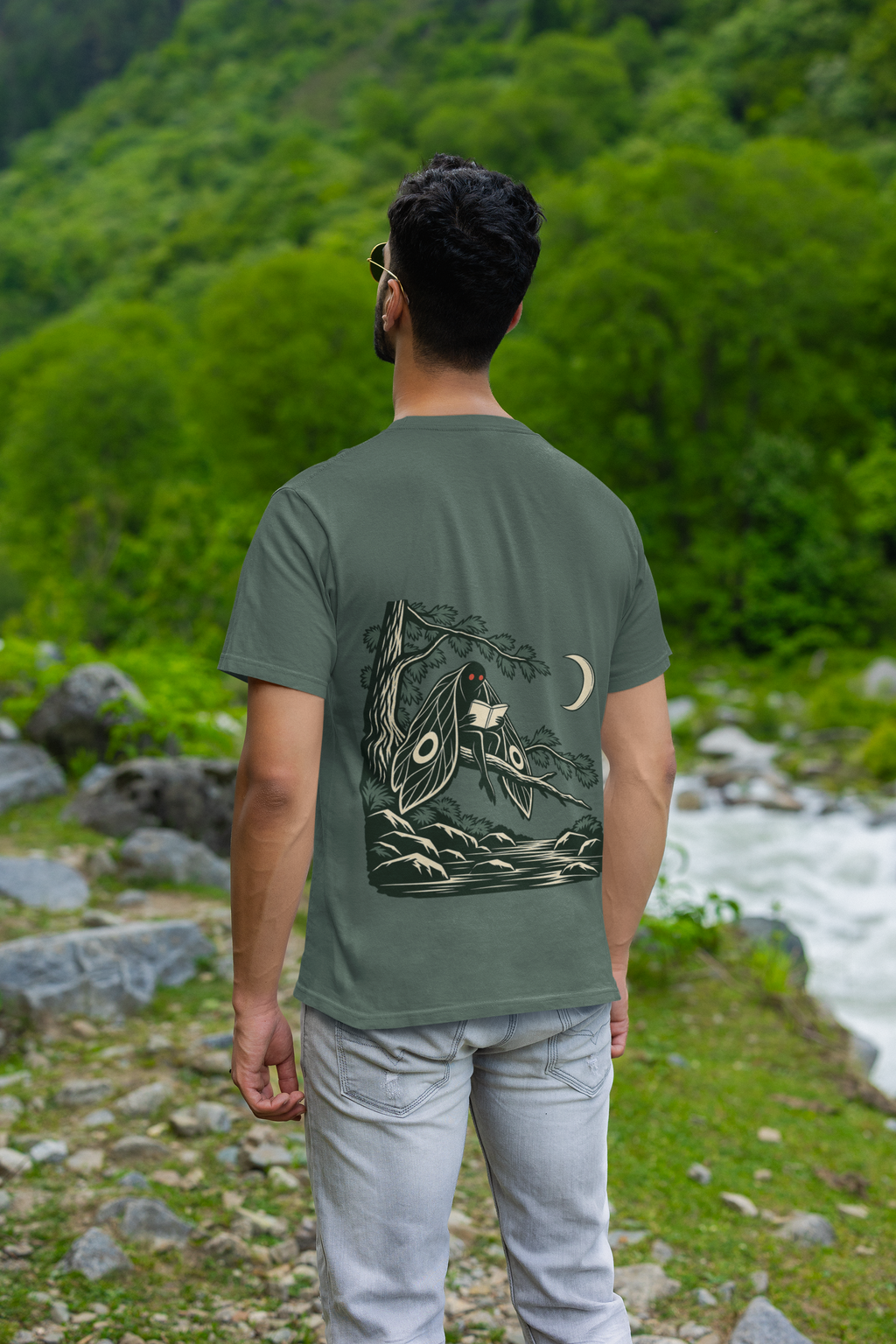Night Library Garment-Dyed Tee in Blue Spruce, worn by a man seen from behind by a mountain river, showing the large print on the back. Digital product mockup.