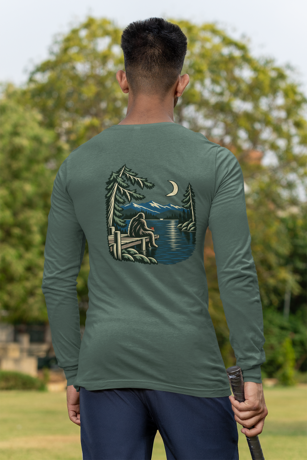Quiet Hours Garment-Dyed Long-Sleeve Tee in Blue Spruce, worn by a man seen from behind at a cricket field, showing the large print on the back. Digital product mockup.