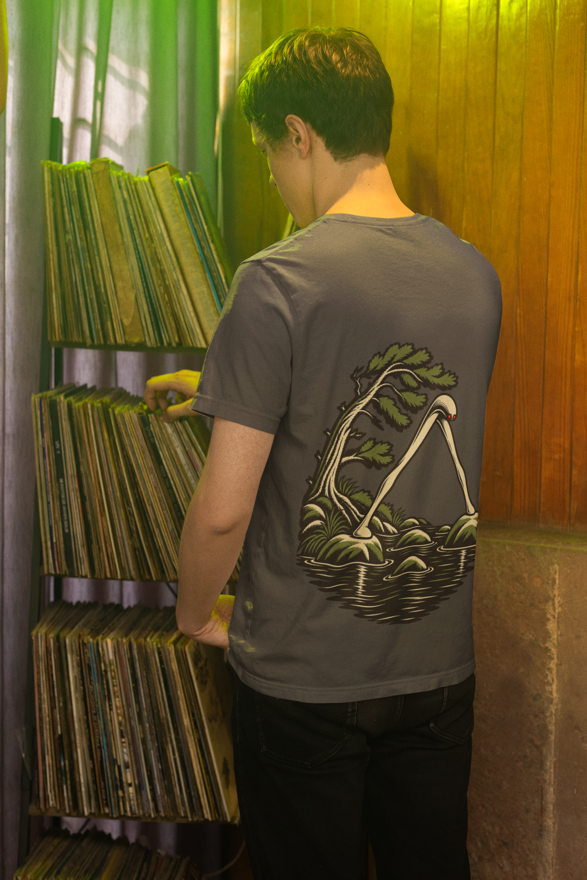 Small Hours Garment-Dyed Tee in Graphite, worn by a man seen from behind browsing a record shop, showing the large print on the back. Digital product mockup.