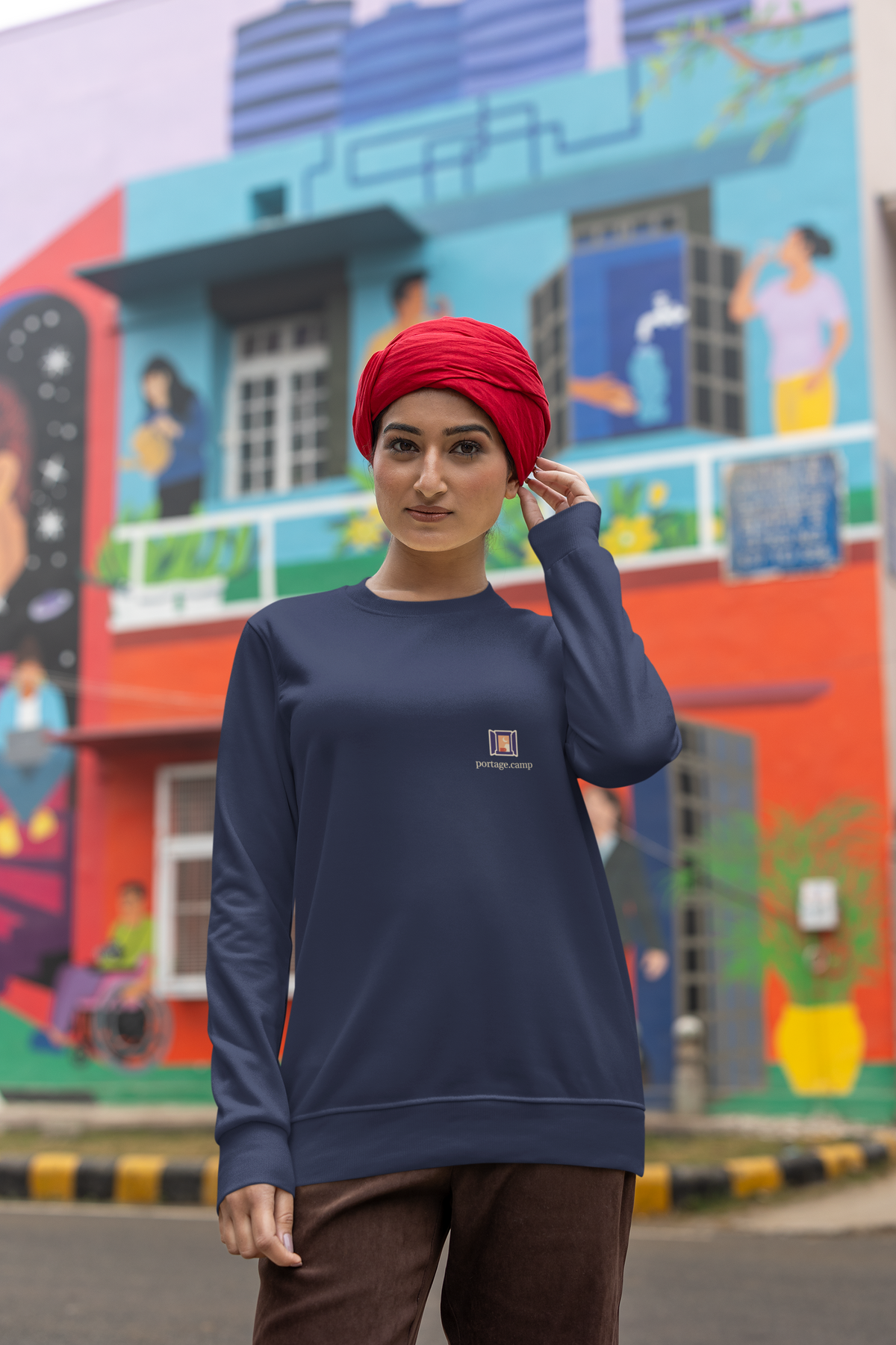 Open Late Garment-Dyed Crewneck in True Navy, worn by a woman in a turban on a busy street, with the Open Late logo and portage.camp on the chest. Digital product mockup.