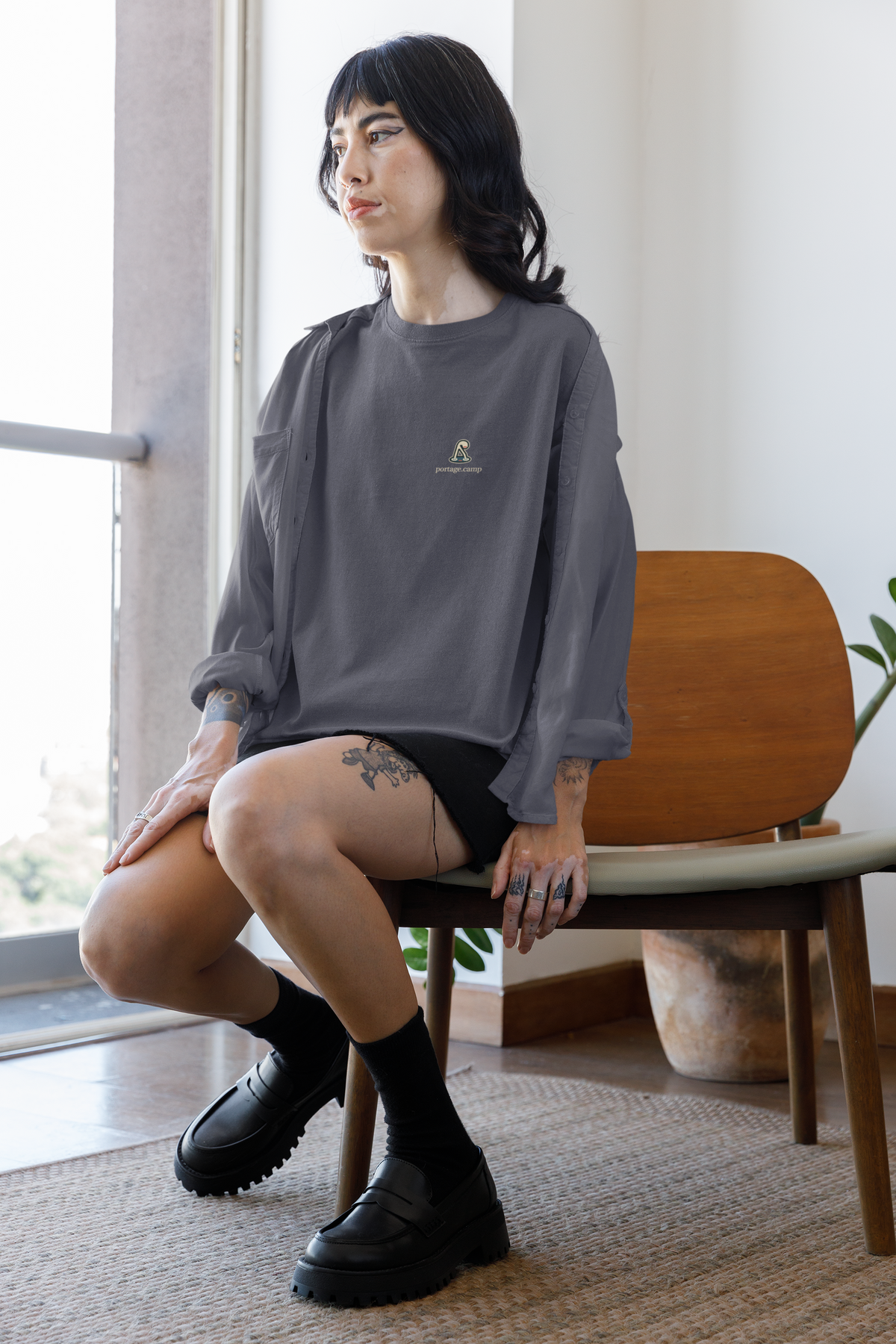 Small Hours Garment-Dyed Tee in Navy, worn by a woman seated by a window at home, with the Small Hours logo and portage.camp on the chest. Digital product mockup.