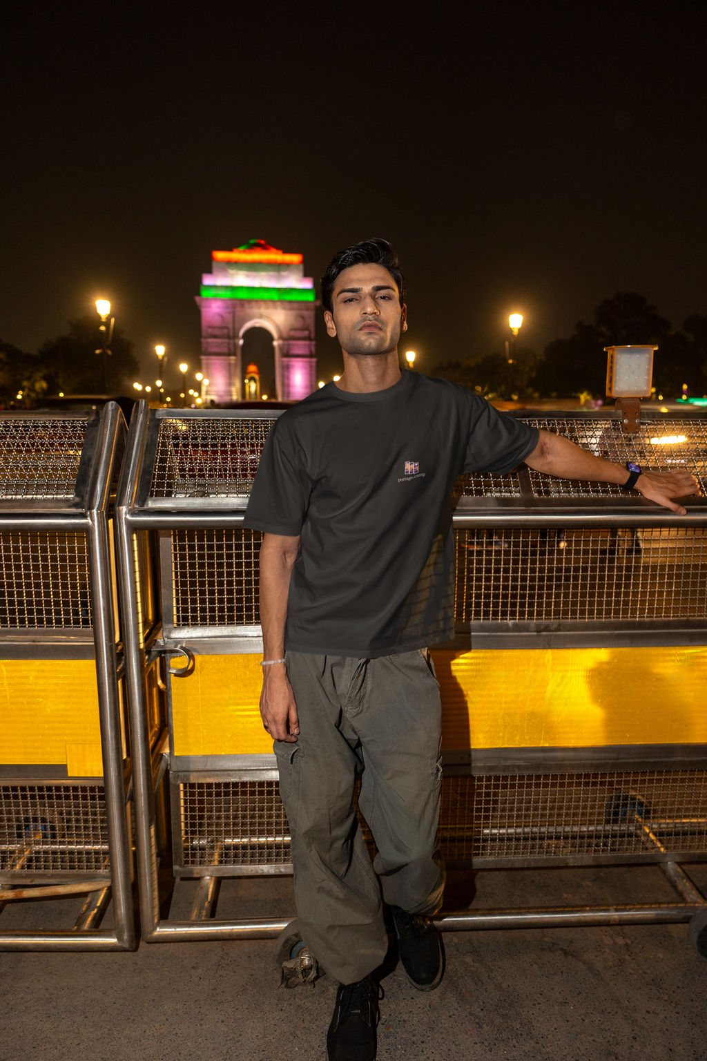 Stay for the Sky Garment-Dyed Tee in Black, worn by a man on a bridge over the city at night, with the Stay for the Sky logo and portage.camp on the chest. Digital product mockup.