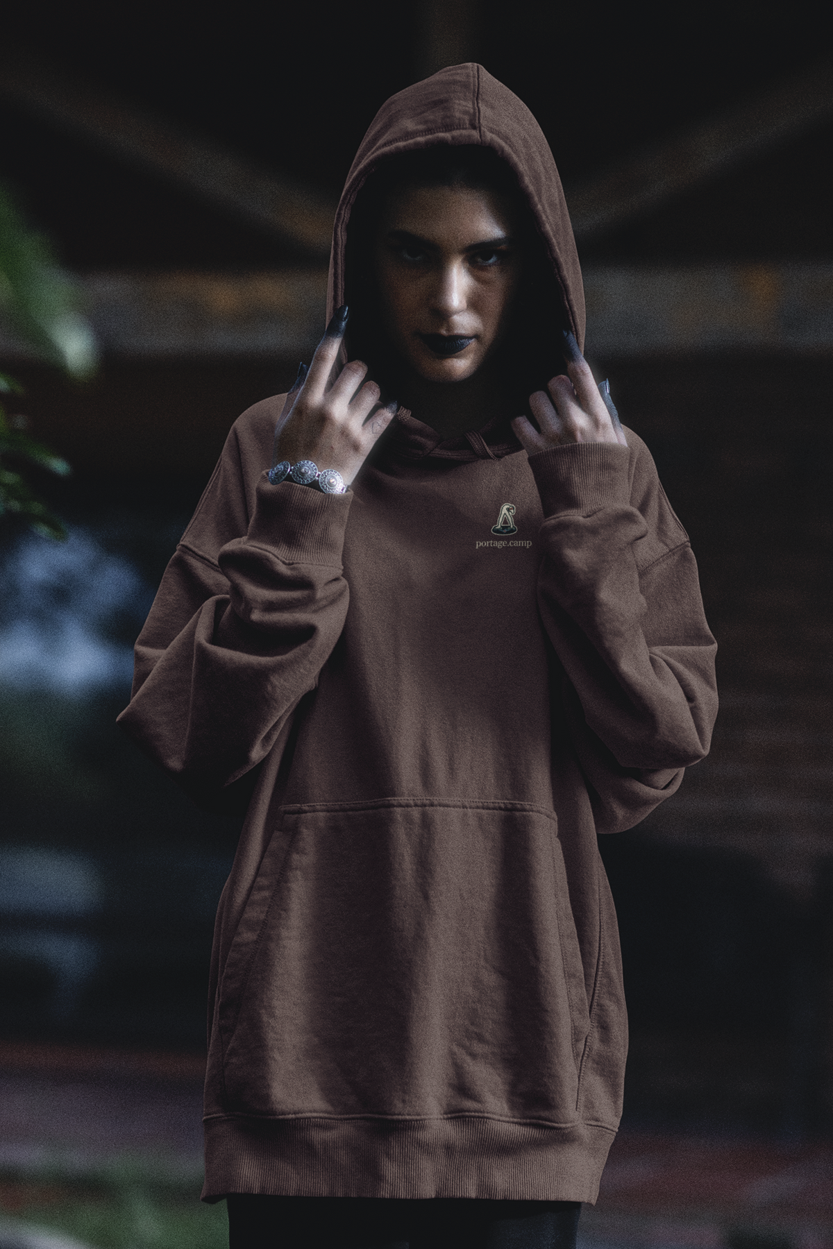 Small Hours Heavyweight Hoodie in Brown, worn by a woman in a dark forest, with the Small Hours logo and portage.camp on the left chest. Digital product mockup.