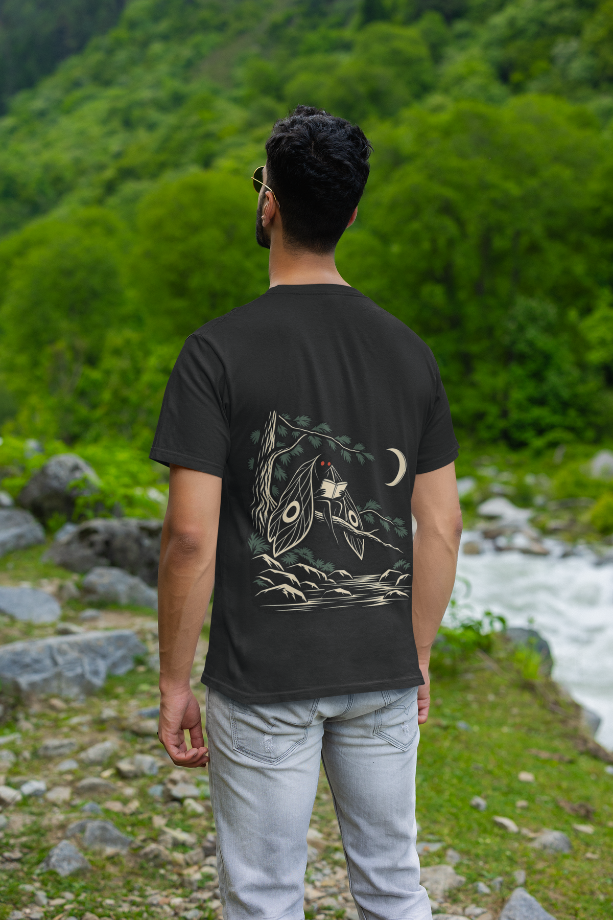 Night Library Garment-Dyed Tee in Black, worn by a man seen from behind by a mountain river, showing the large print on the back. Digital product mockup.