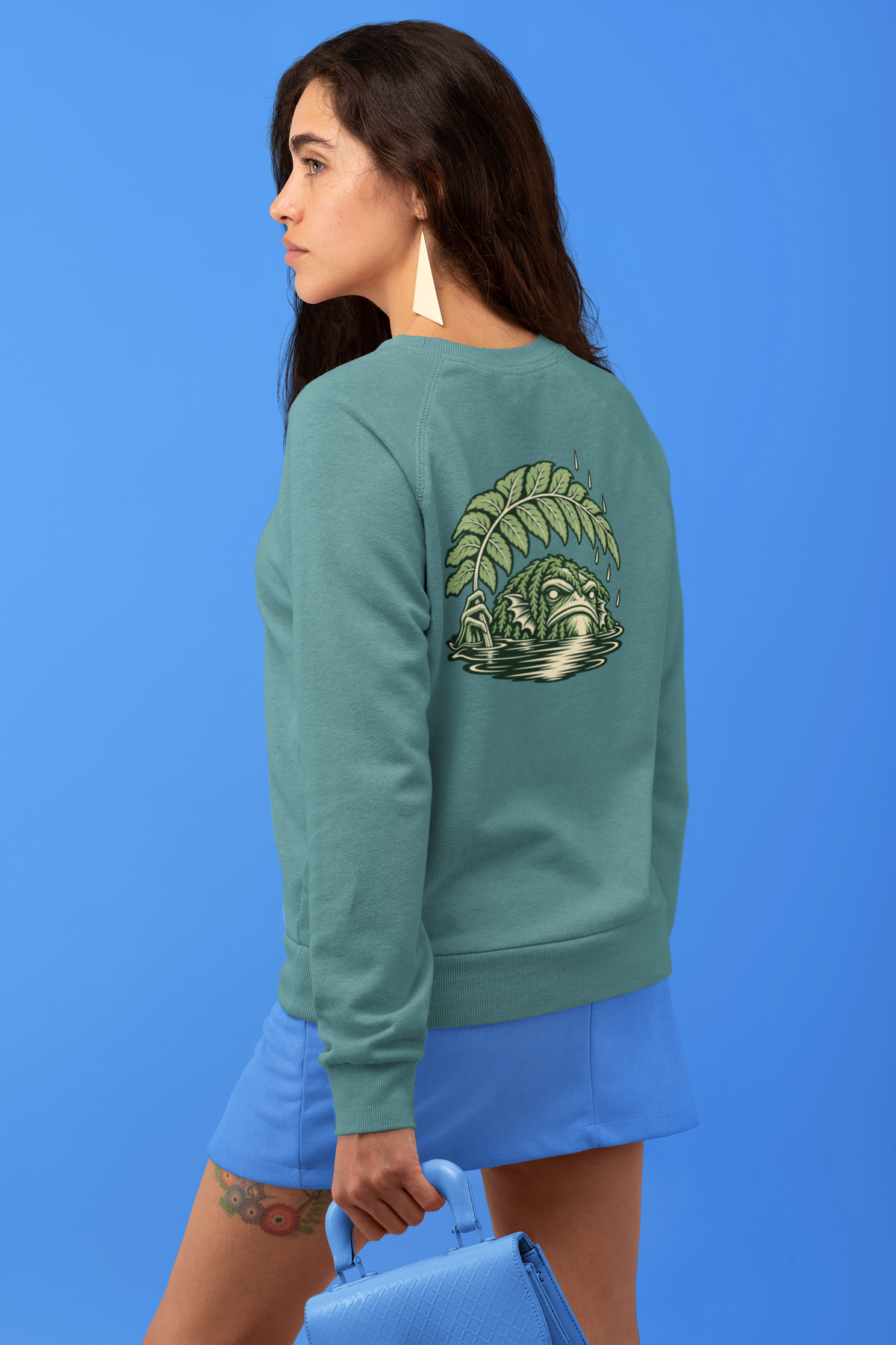 Rain Check Garment-Dyed Crewneck in Blue Spruce, worn by a woman seen from behind against a blue backdrop, showing the large print on the back. Digital product mockup.