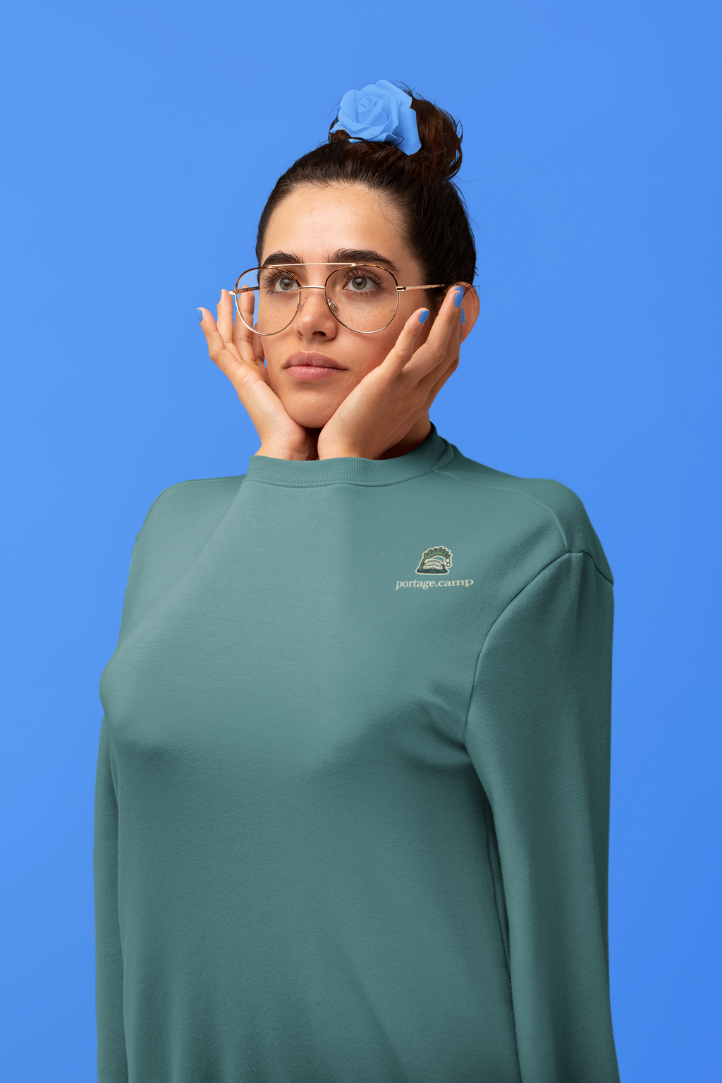 Rain Check Garment-Dyed Crewneck in Blue Spruce, worn by a woman in a studio, arms tucked inside the sleeves, with the Rain Check logo and portage.camp on the chest. Digital product mockup.