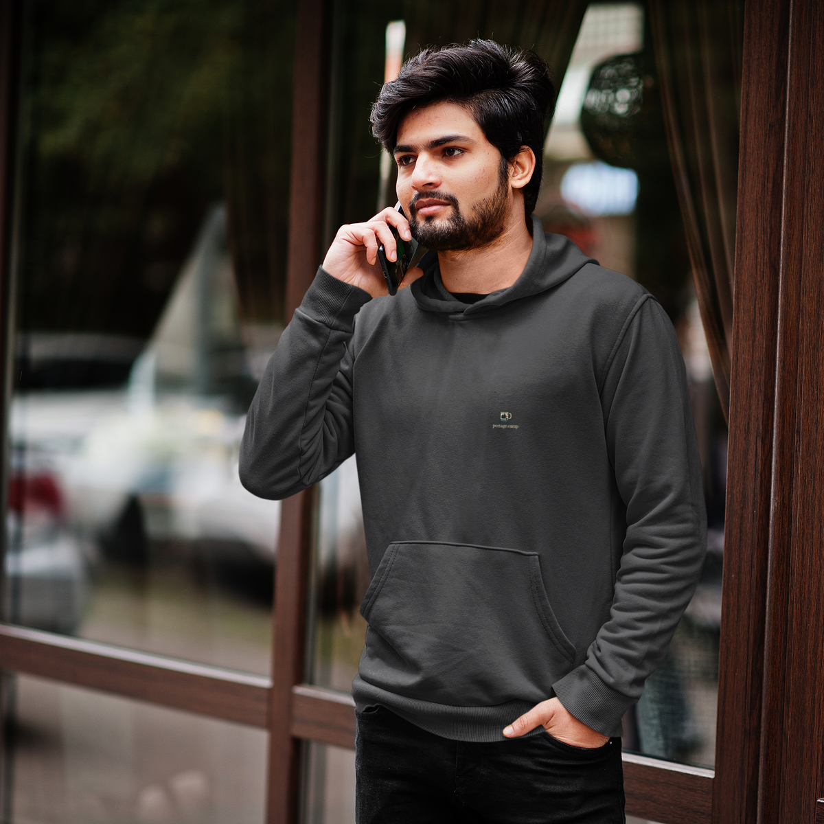 Quiet Hours Heavyweight Hoodie in Black, worn by a man by a café window, on a phone call, with the Quiet Hours logo and portage.camp on the left chest. Digital product mockup.