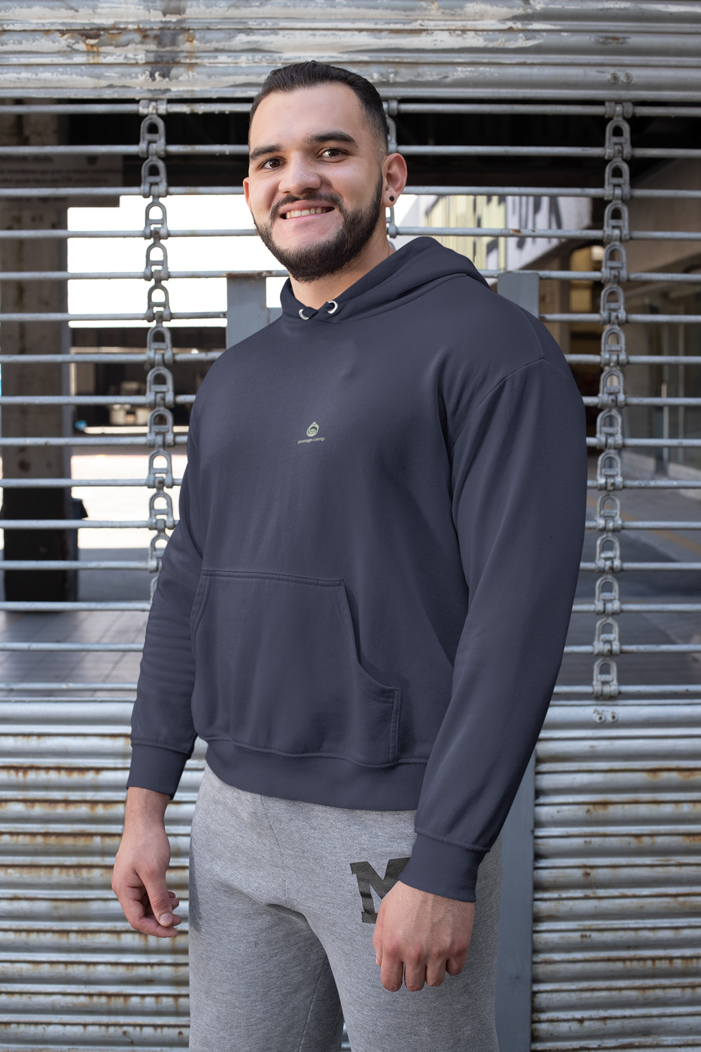 Rain Check Midweight Hoodie in Classic Navy, worn by a man against a metal fence, with the Rain Check logo and portage.camp on the chest. Digital product mockup.