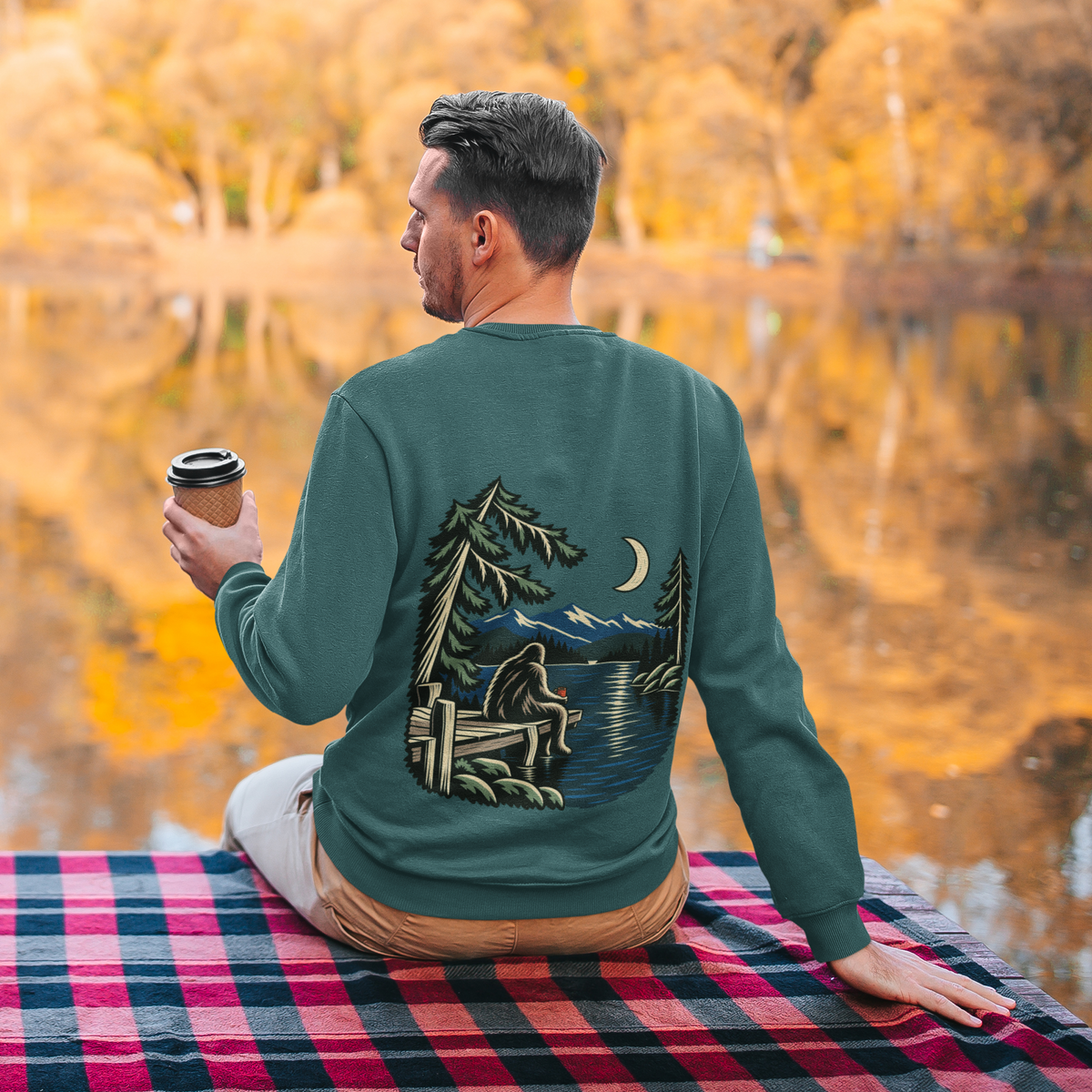 Quiet Hours Garment-Dyed Crewneck in Blue Spruce, worn by a man seen from behind seated by a lake in autumn, showing the large print on the back. Digital product mockup.