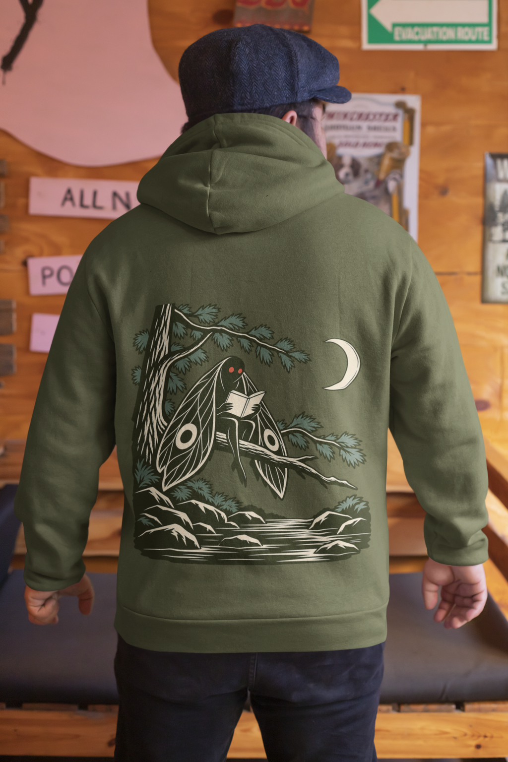 Night Library Midweight Hoodie in Army, worn by a man seen from behind in a wood-panelled bar, showing the large print on the back. Digital product mockup.