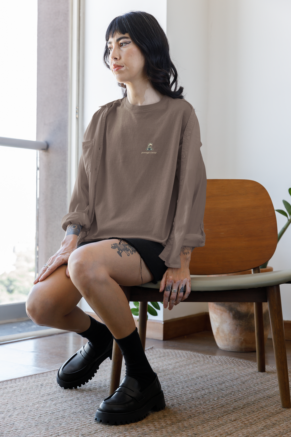 Small Hours Garment-Dyed Tee in Espresso, worn by a woman seated by a window at home, with the Small Hours logo and portage.camp on the chest. Digital product mockup.