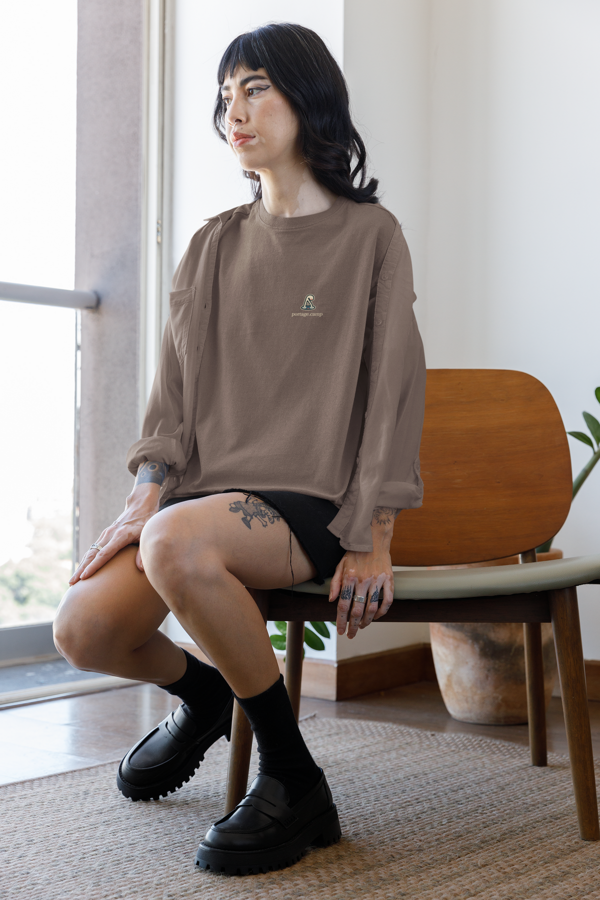 Small Hours Garment-Dyed Tee in Espresso, worn by a woman seated by a window at home, with the Small Hours logo and portage.camp on the chest. Digital product mockup.