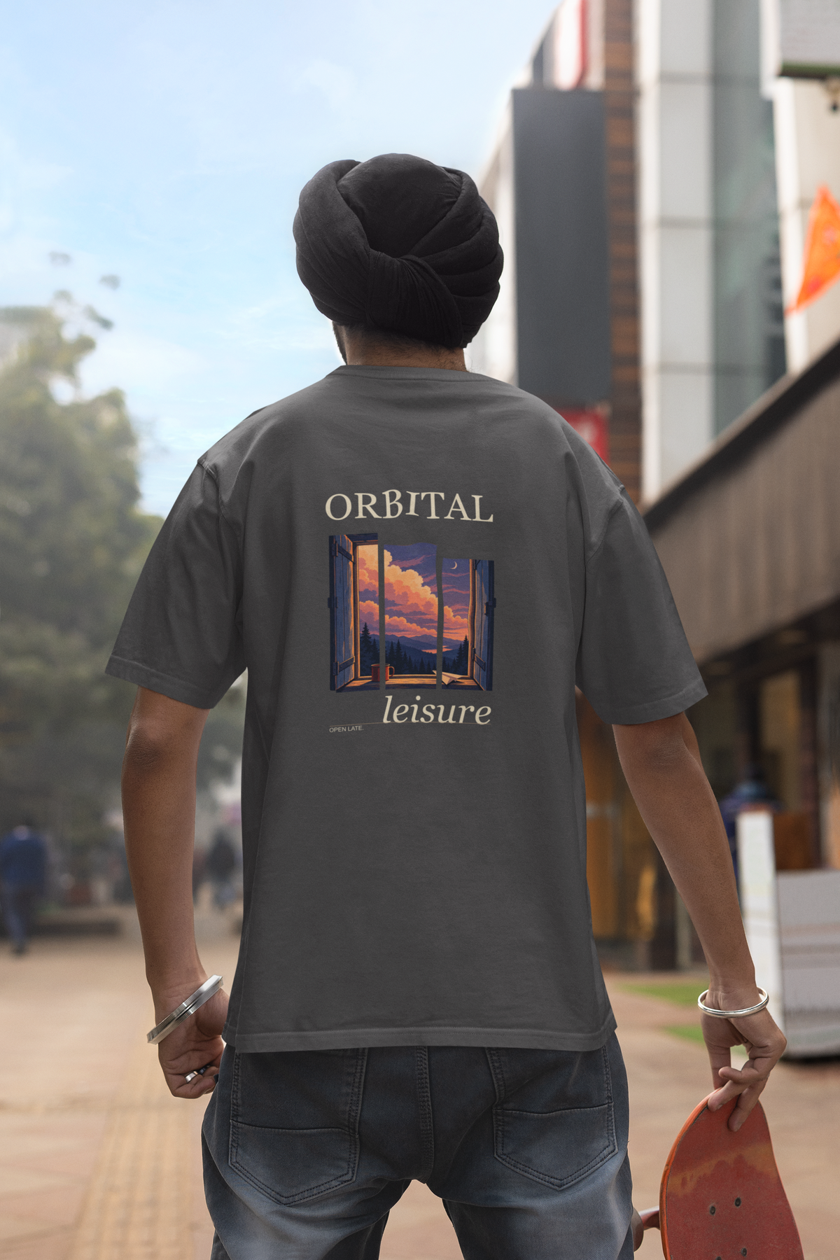 Open Late Garment-Dyed Tee in Graphite, worn by a man seen from behind in a turban on a busy street, showing the large print on the back. Digital product mockup.