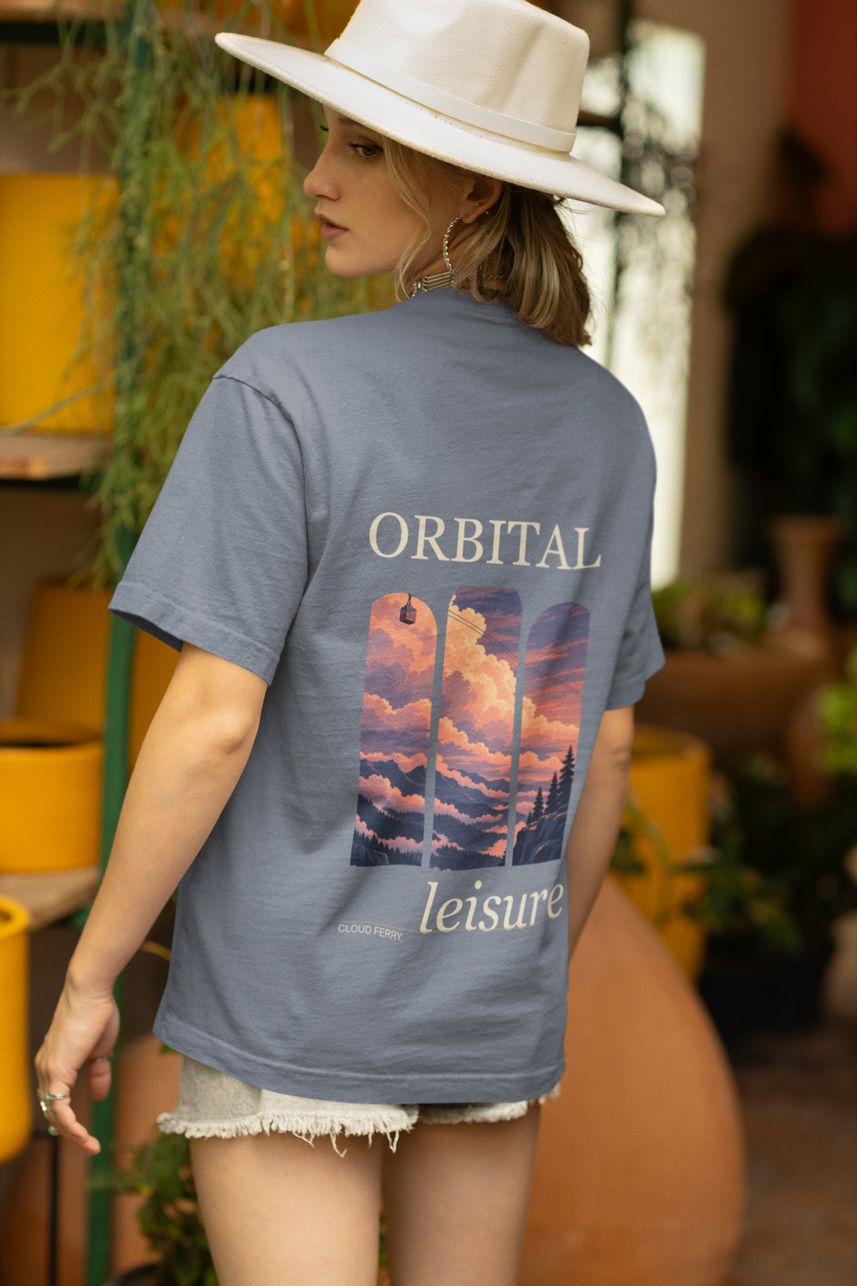 Cloud Ferry Garment-Dyed Tee in Blue Jean, worn by a woman seen from behind in a sun hat among potted plants, showing the large print on the back. Digital product mockup.