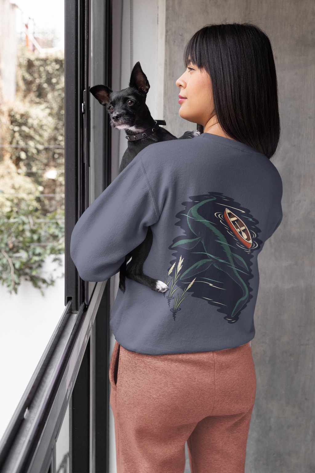 Seen Briefly Garment-Dyed Crewneck in Denim, worn by a woman seen from behind at home, holding her dog, showing the large print on the back. Digital product mockup.