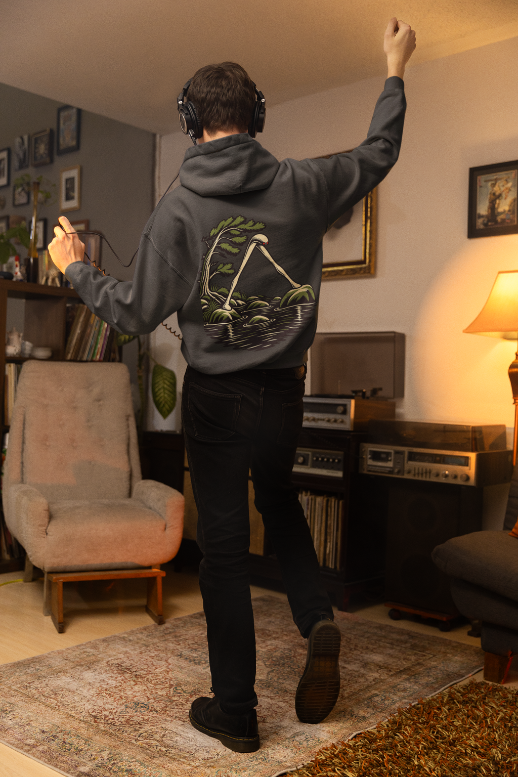 Small Hours Heavyweight Hoodie in Black, worn by a man seen from behind in a living room at night, showing the large print on the back. Digital product mockup.