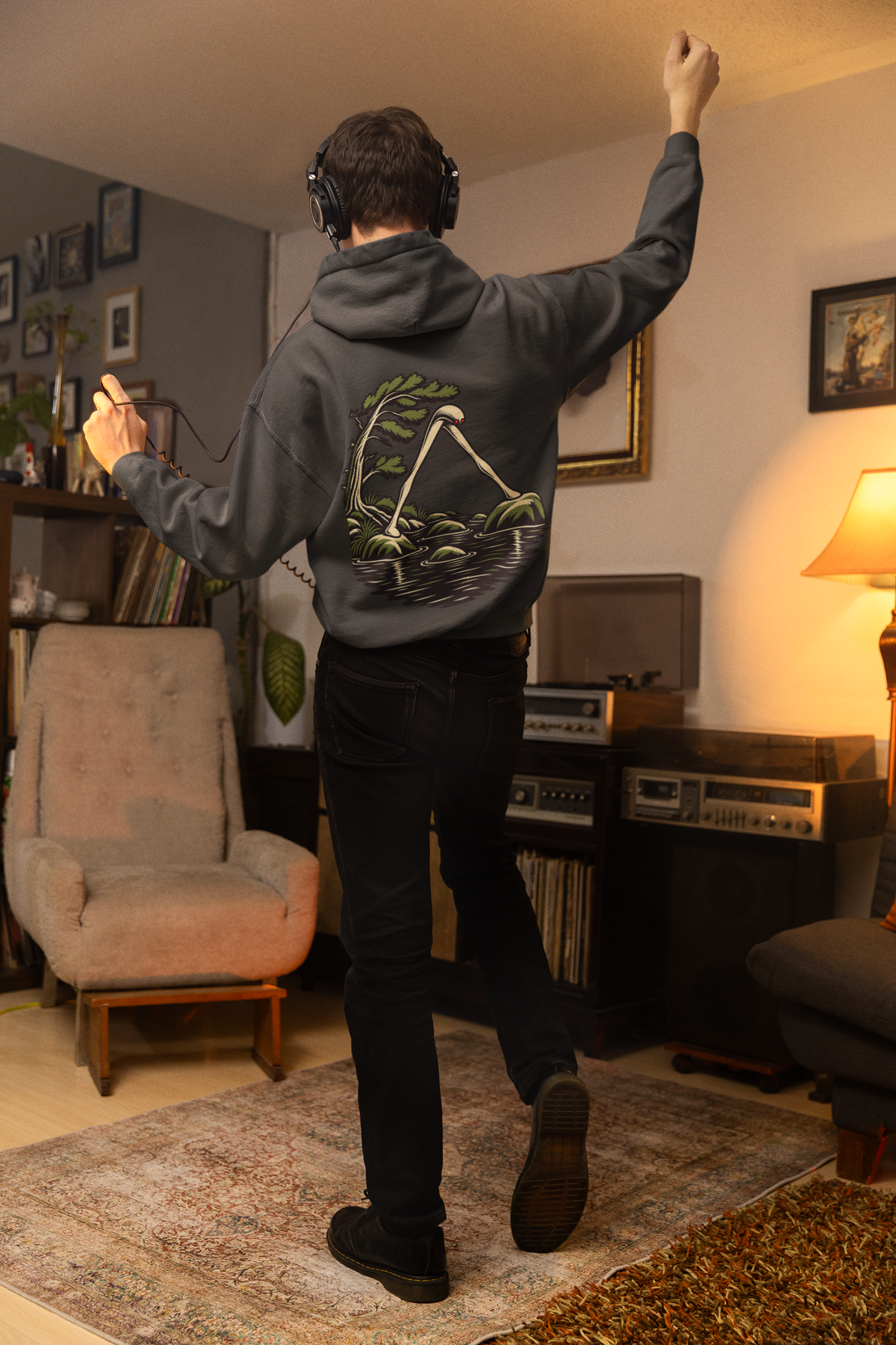 Small Hours Heavyweight Hoodie in Black, worn by a man seen from behind in a living room at night, showing the large print on the back. Digital product mockup.