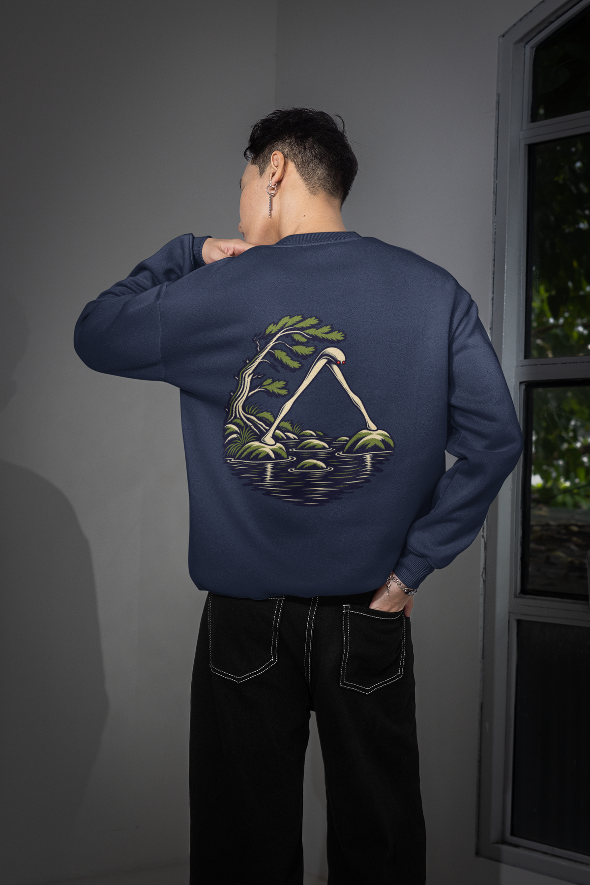 Small Hours Garment-Dyed Crewneck in True Navy, worn by a man seen from behind in a room by a window, showing the large print on the back. Digital product mockup.
