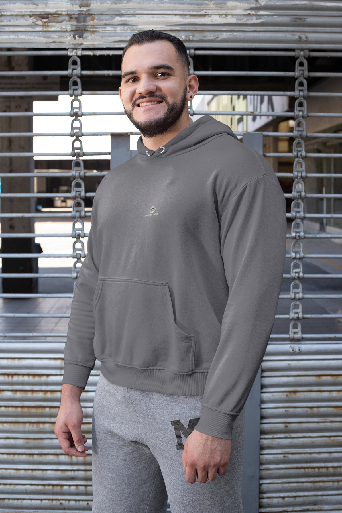 Rain Check Midweight Hoodie in Charcoal, worn by a man against a metal fence, with the Rain Check logo and portage.camp on the chest. Digital product mockup.