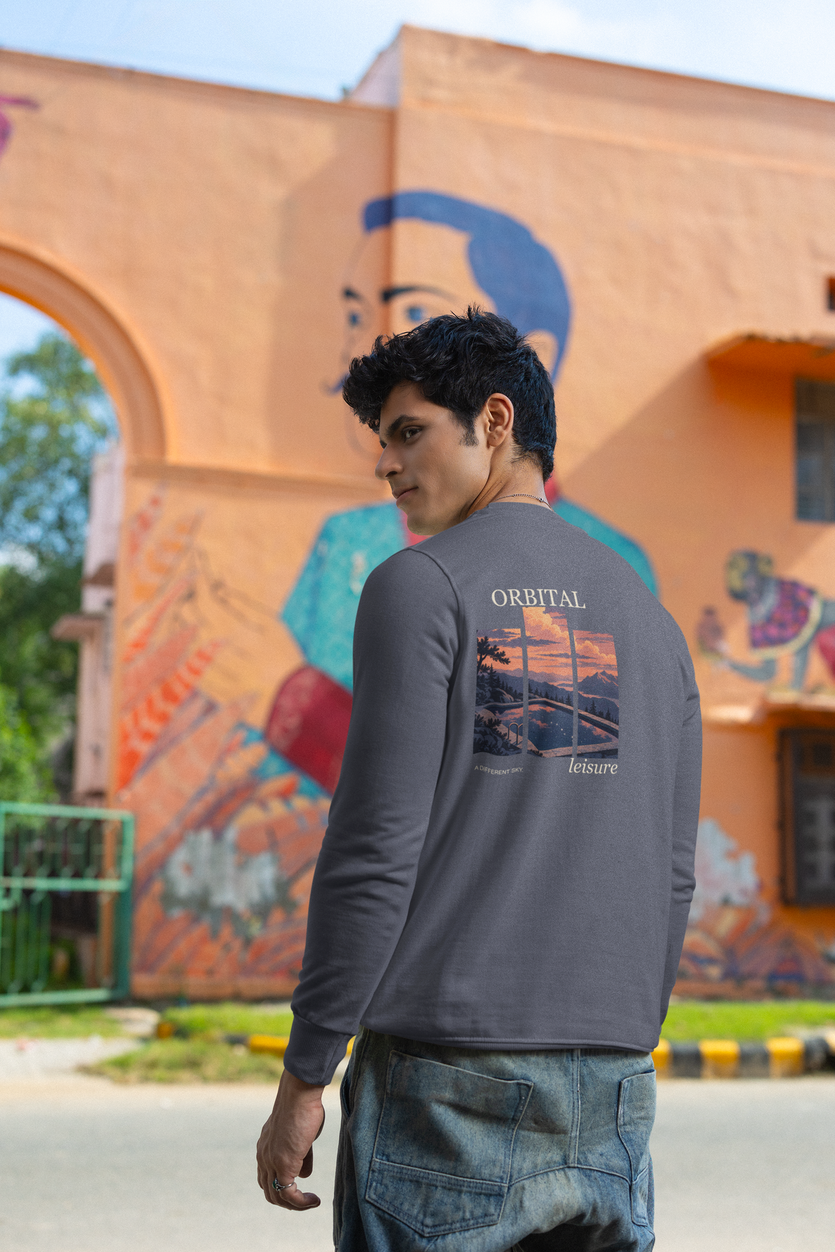 A Different Sky Garment-Dyed Crewneck in Denim, worn by a man seen from behind by a painted wall in the city, showing the large print on the back. Digital product mockup.