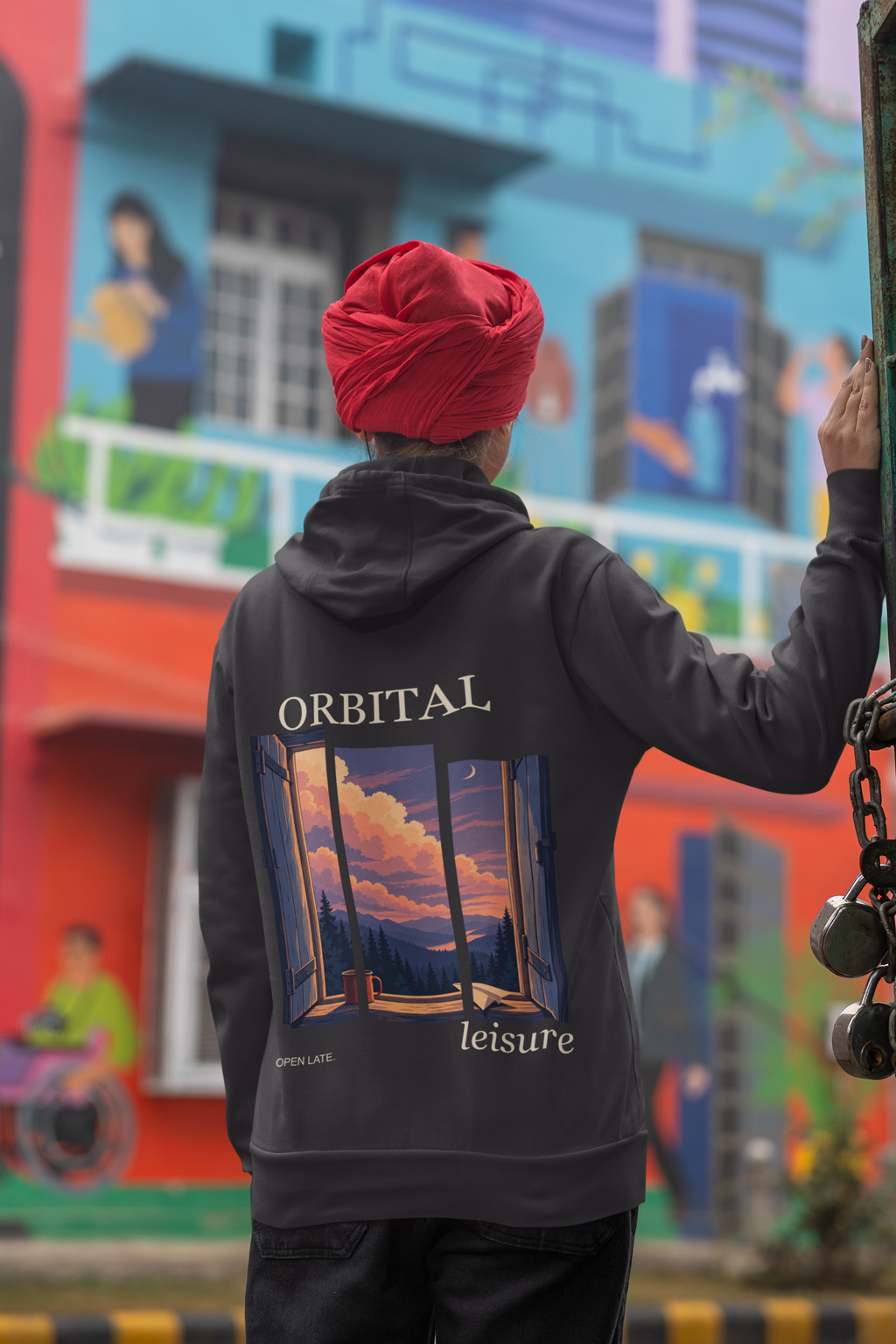 Open Late Zip Hoodie in Black, worn by a woman seen from behind in a turban by a painted mural, showing the large print on the back. Digital product mockup.
