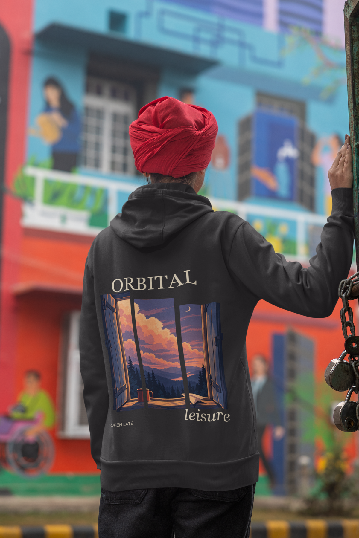 Open Late Zip Hoodie in Black, worn by a woman seen from behind in a turban by a painted mural, showing the large print on the back. Digital product mockup.