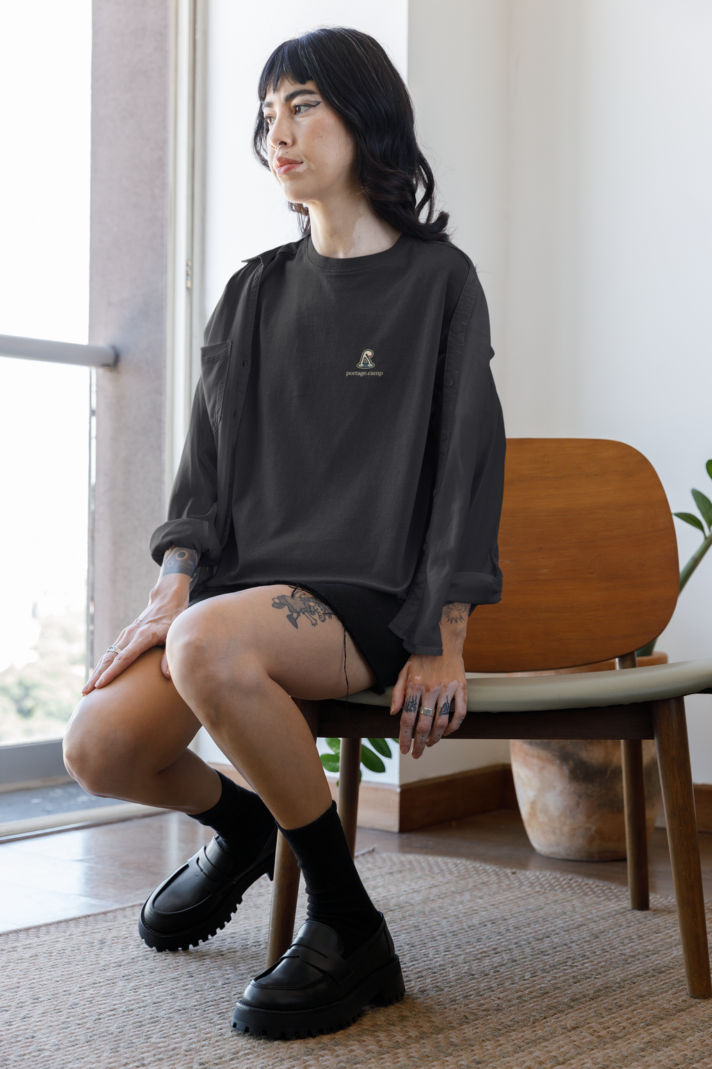 Small Hours Garment-Dyed Tee in Black, worn by a woman seated by a window at home, with the Small Hours logo and portage.camp on the chest. Digital product mockup.