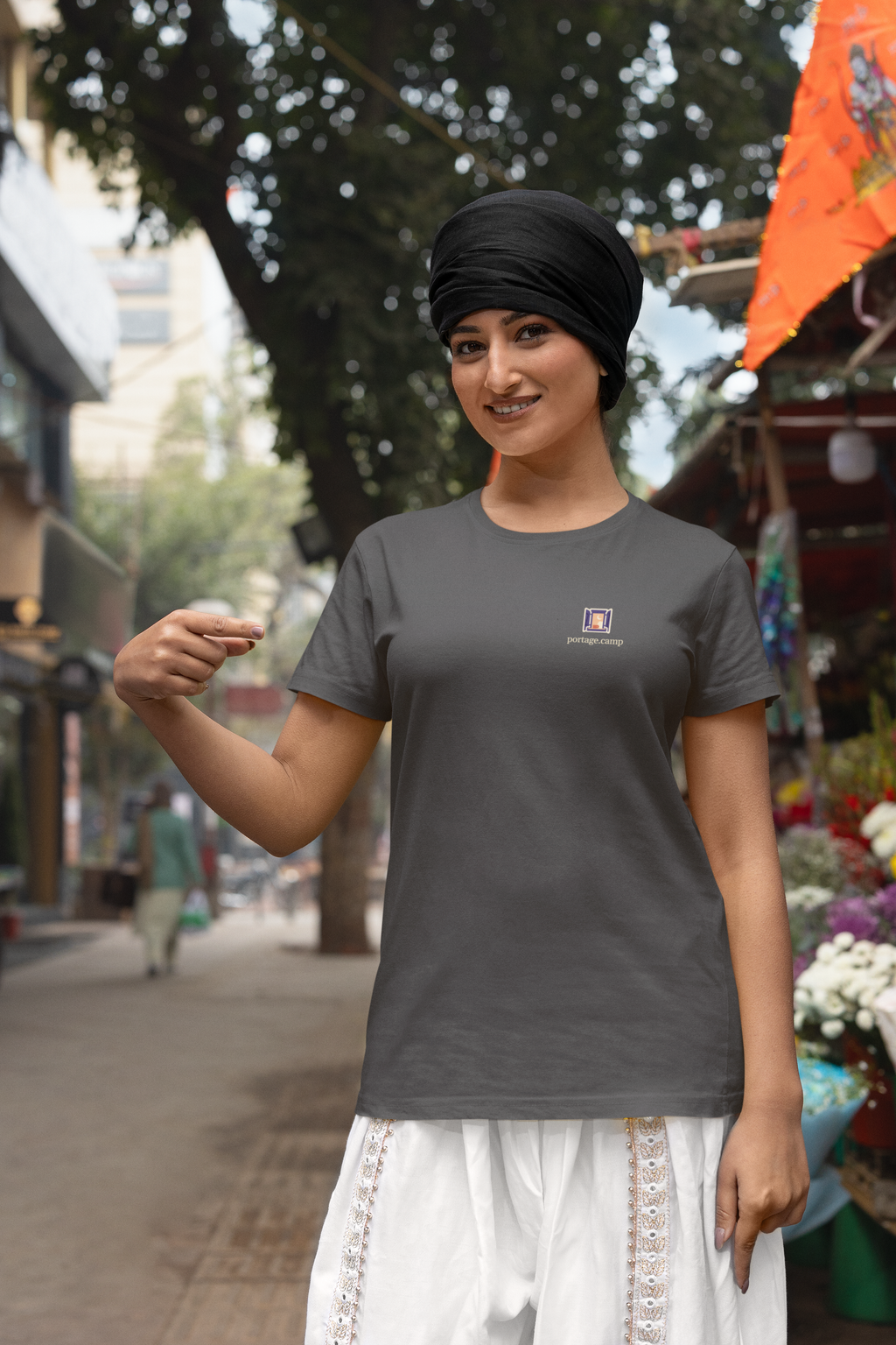 Open Late Garment-Dyed Tee in Graphite, worn by a woman in a turban by a flower market, with the Open Late logo and portage.camp on the chest. Digital product mockup.